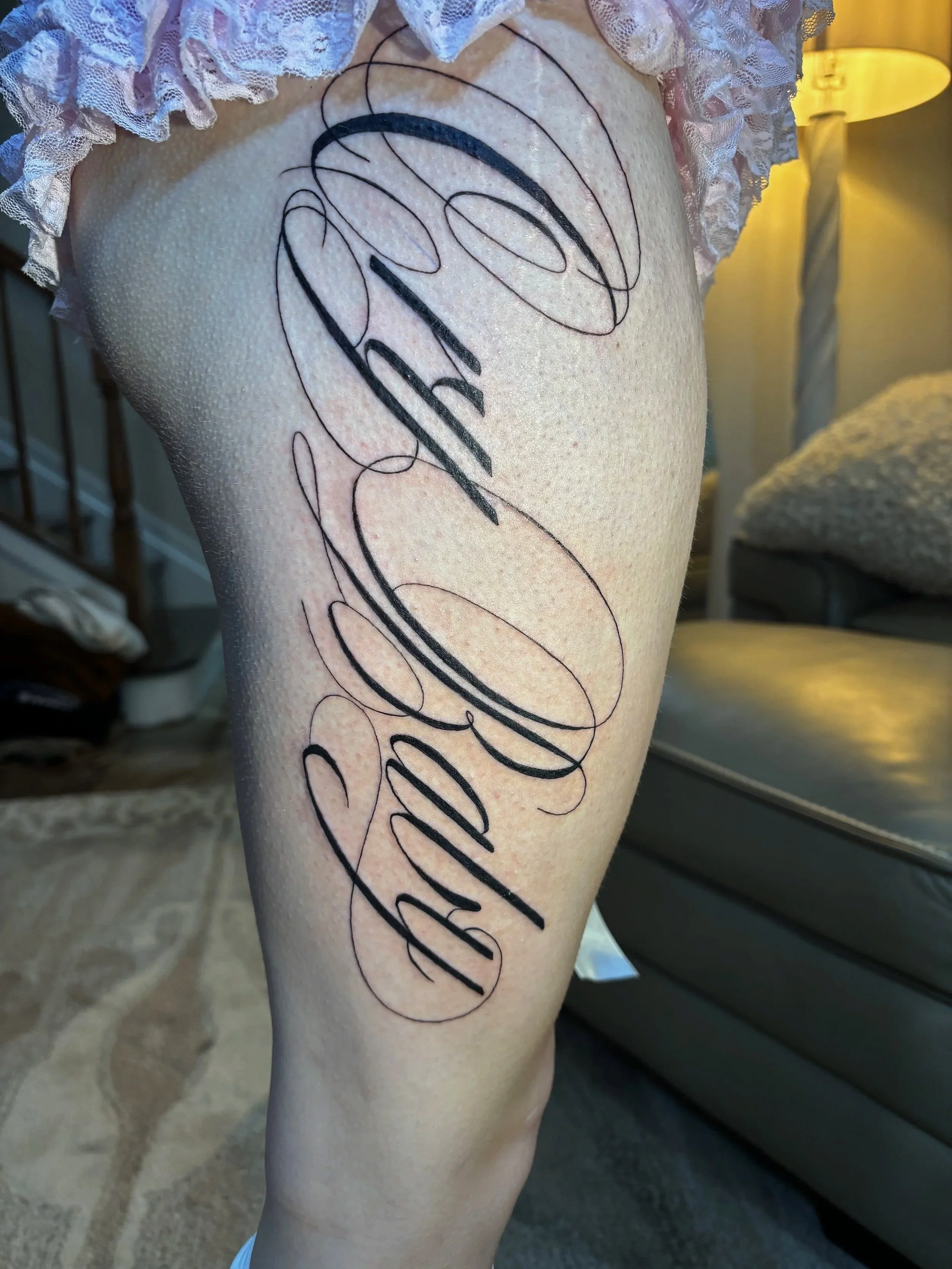 Close-up of a person's thigh with a fresh, black tattoo of cursive lettering that reads 'cry baby'. The person is wearing light pink, ruffled lace underwear, and the background shows a living room with a beige carpet, a black couch, and a white lamp.