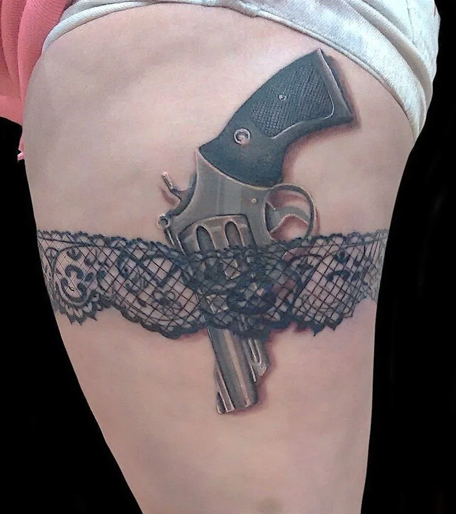 Tattoo of a revolver gun with a black handle, positioned specific orientation, underneath a black fishnet tattoo pattern, on a person's upper thigh.