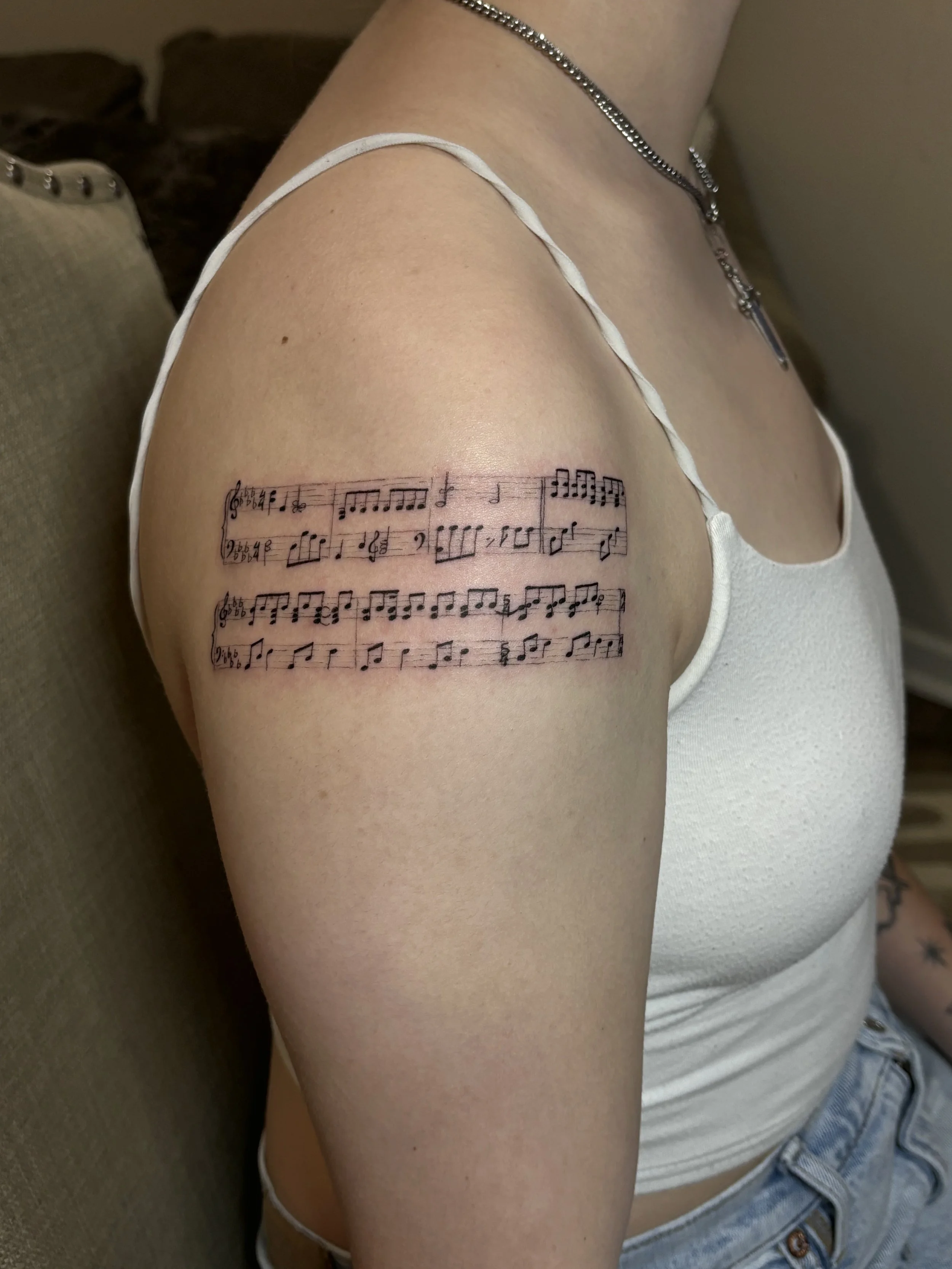 A woman's upper arm with a tattoo of sheet music, wearing a white top and a necklace, sitting on a beige couch.