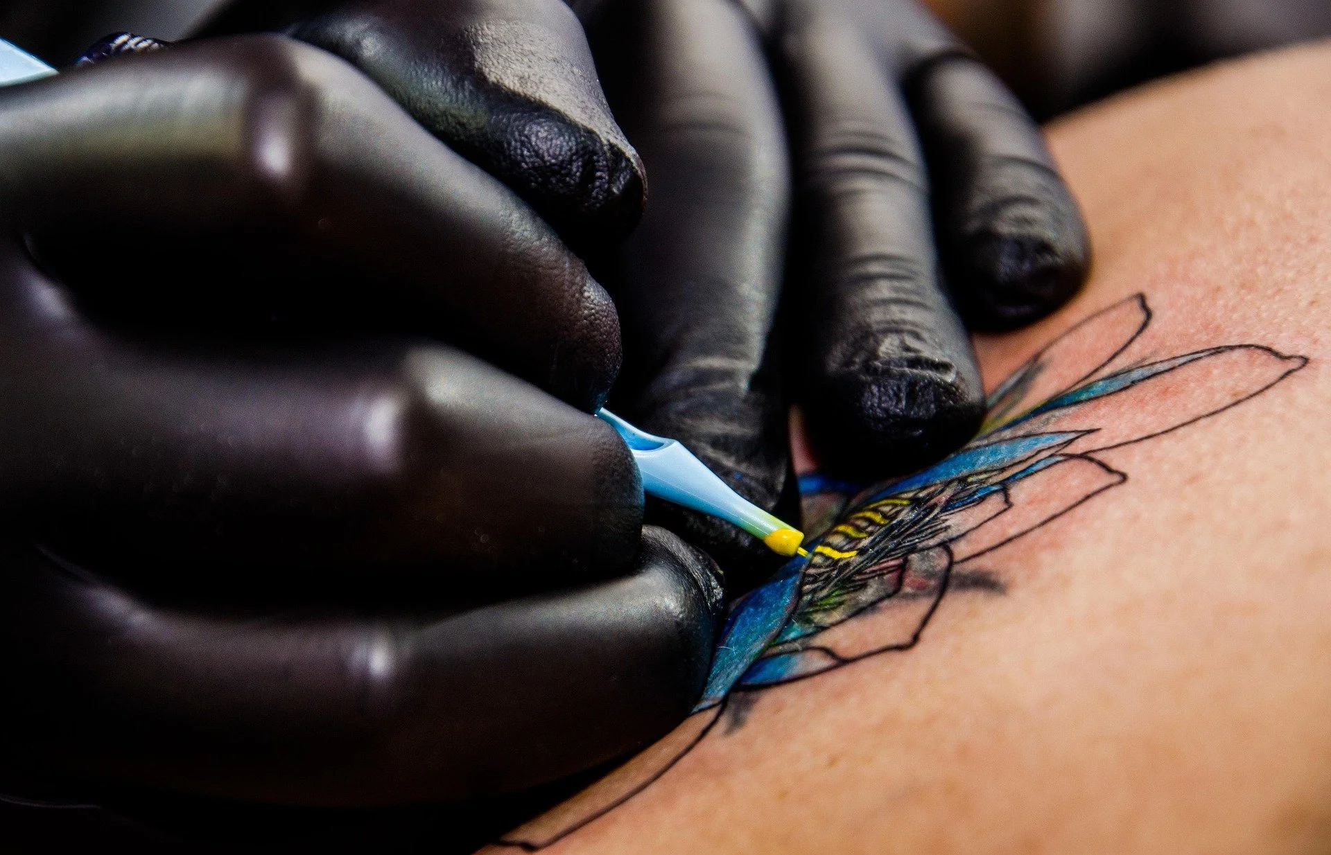 A person wearing black gloves is inking a colorful butterfly tattoo on their skin with a tattoo machine.
