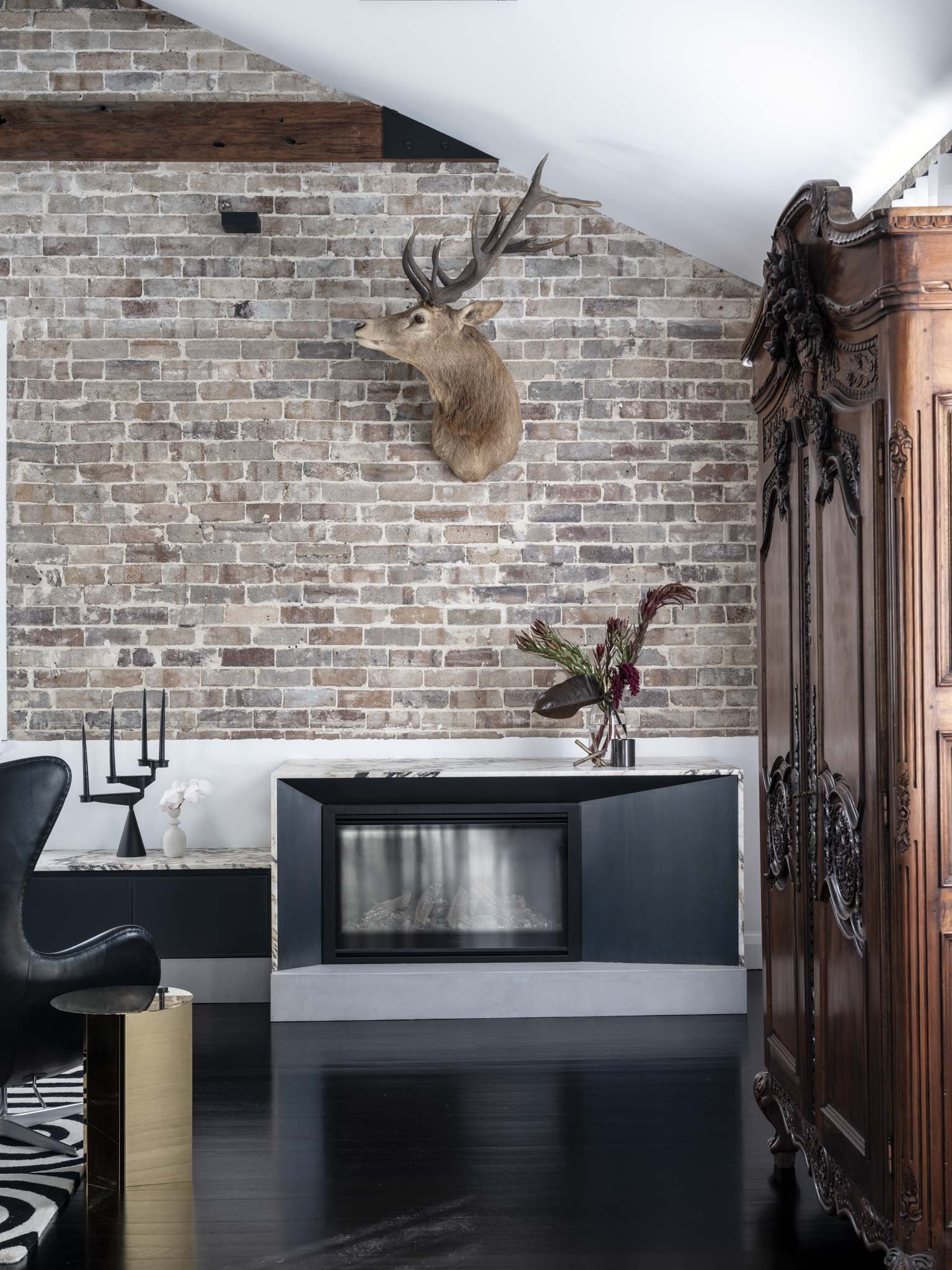 Interior of a living room with a brick wall, a mounted deer head, a stylish fireplace, an ornate wooden cabinet, a black armchair, and decorative items including a vase with flowers and candles.