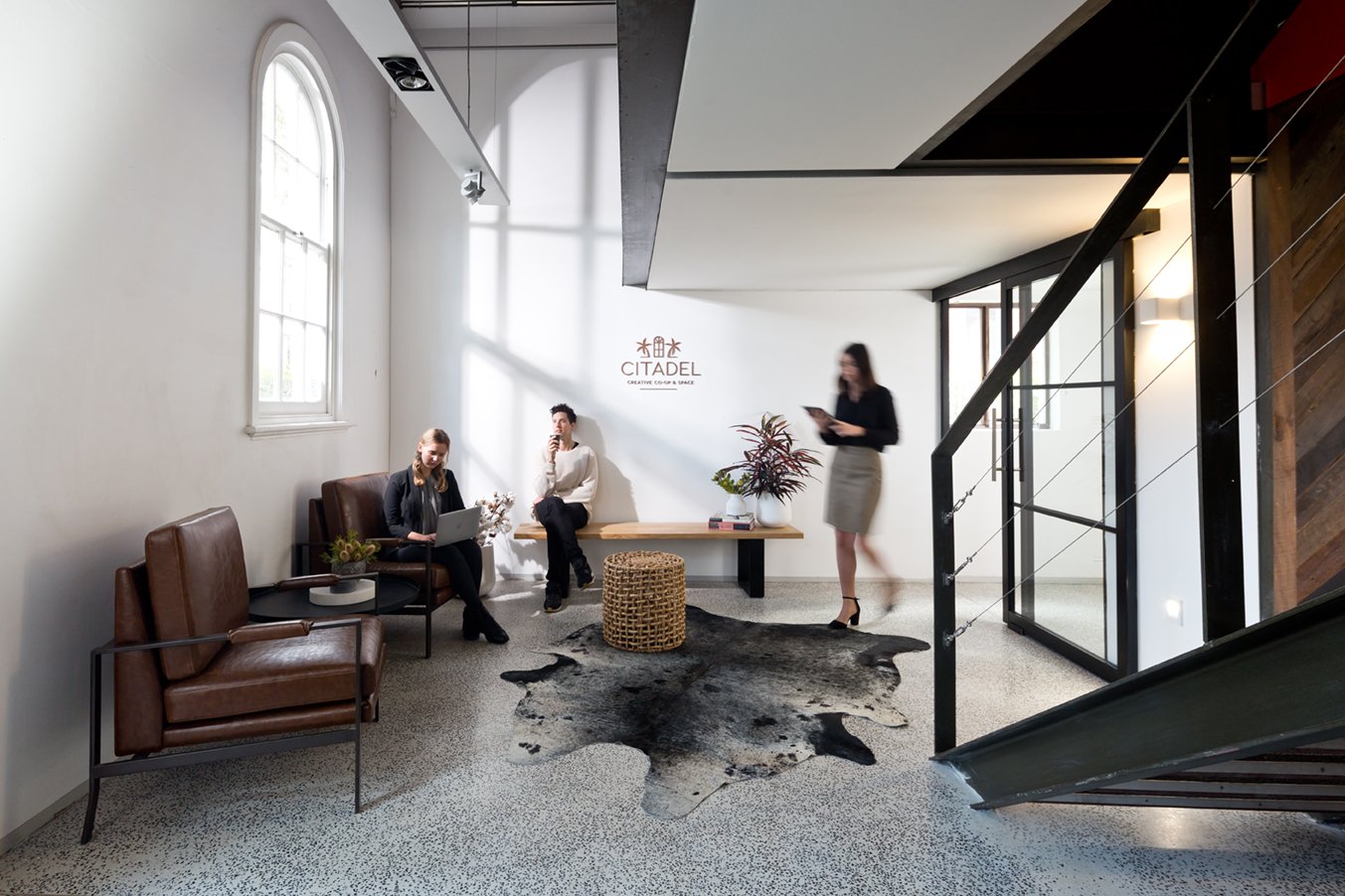 Modern office lobby with white walls, large arched window, leather chairs, a bench, plants, a cowhide rug, and people using laptops and smartphones