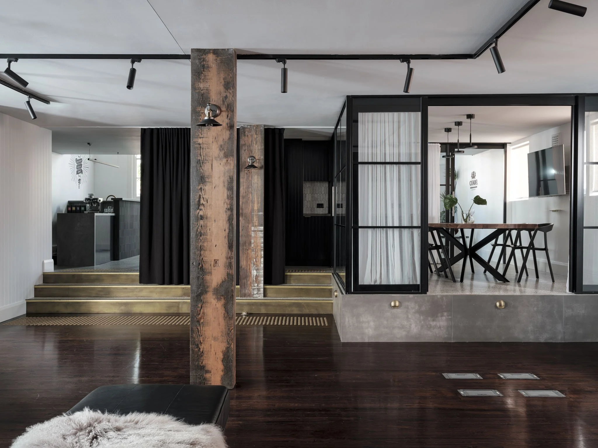 Interior of a modern living space with a dark wooden floor, a black leather ottoman with a white fur throw, a prominent rustic wooden support beam, a stepped platform area with dark curtains, and a dining area with a wooden table and black chairs sep