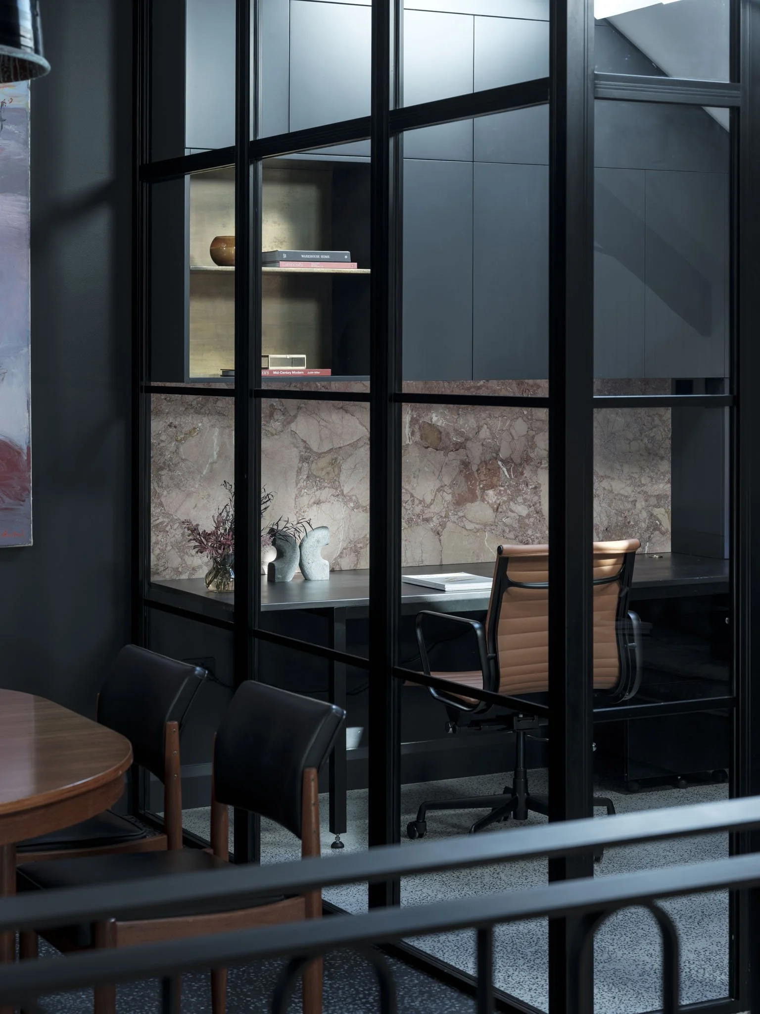 Modern office space with a glass partition, a desk, a tan rolling chair, and a decorative shelf with books and art.