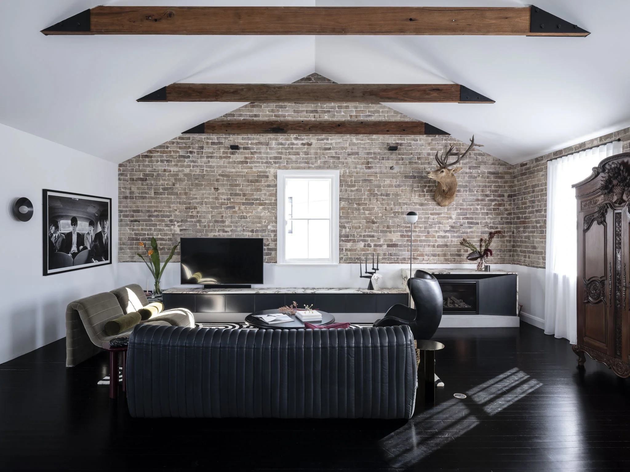 Living room with exposed brick wall, window, mounted deer head, black TV, antique wooden armoire, modern black and beige furniture, and decorative plants.