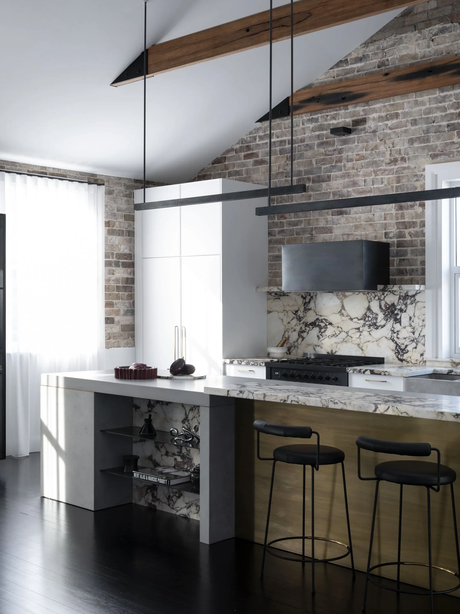 Modern kitchen with exposed brick wall, white cabinetry, marble countertops, hanging black light fixtures, and black bar stools.