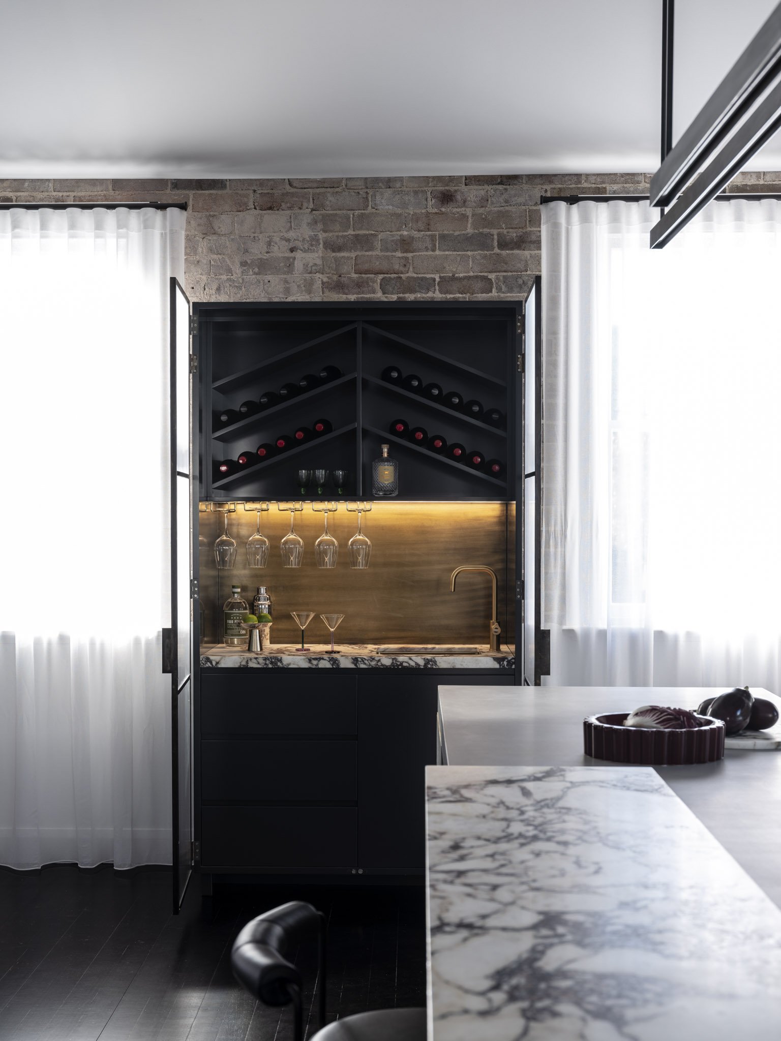Modern kitchen with black cabinets, marble countertop, and a built-in wine storage and glass rack, set against a brick wall with white curtains.