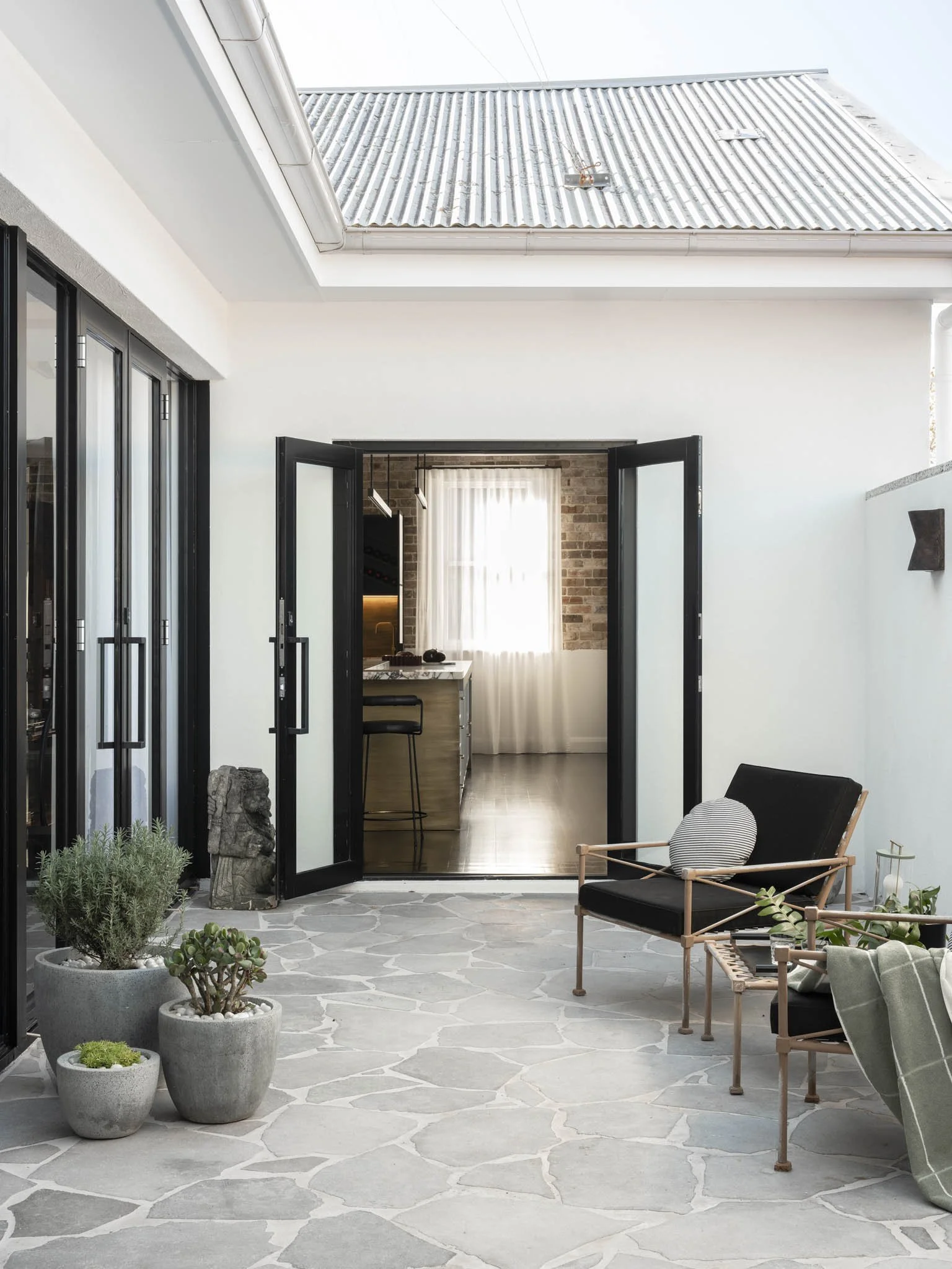 Outdoor patio with stone flooring, black-framed glass doors, potted plants, and outdoor seating with black cushions and wooden frames. The patio area opens into a kitchen with brick wall, white curtains, and black and wooden cabinets.
