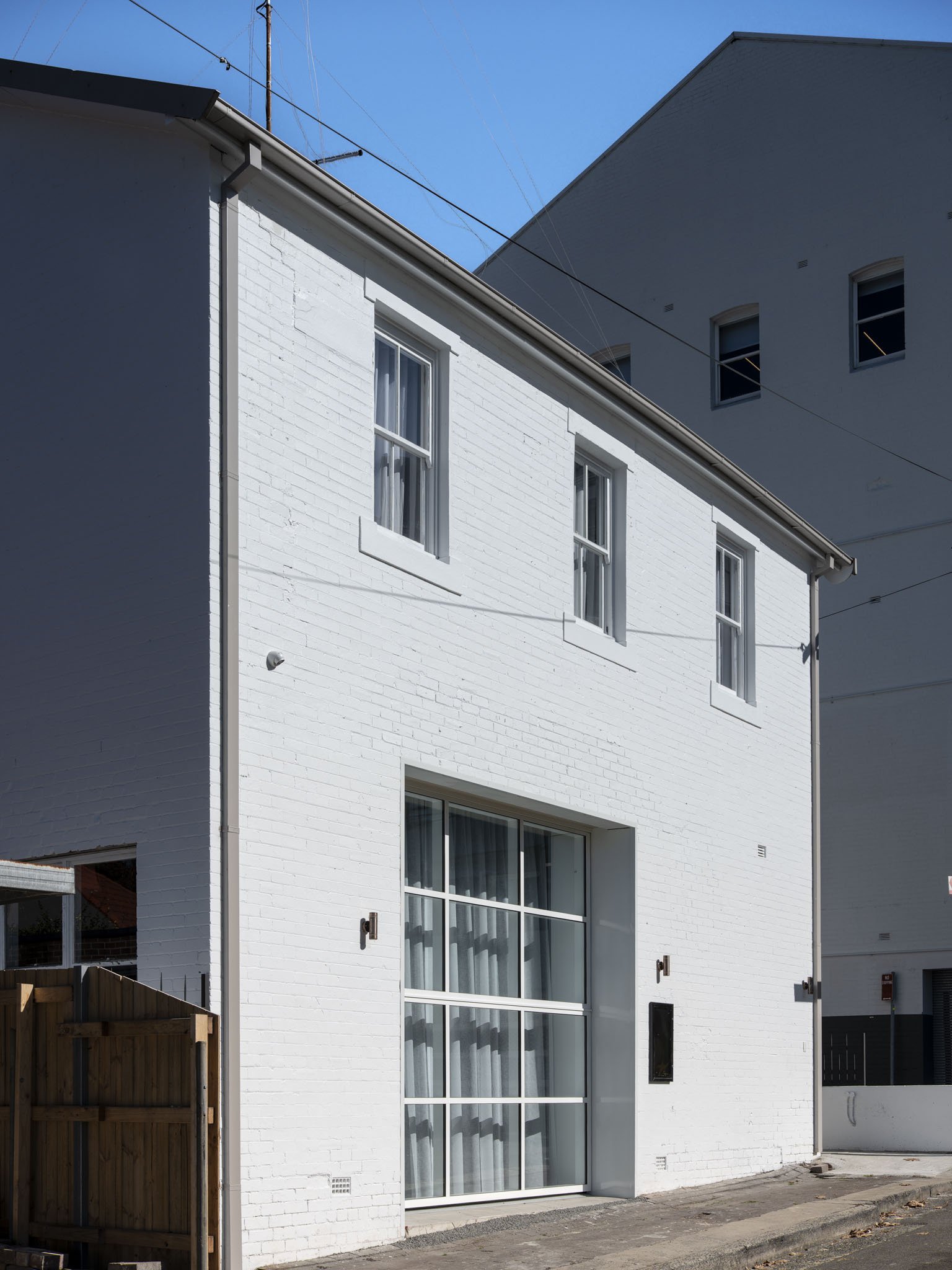 A white brick two-story building with three windows on the upper floor and a large glass door on the lower floor, with curtains visible through the glass. The building is under a clear blue sky, with electrical wires overhead.