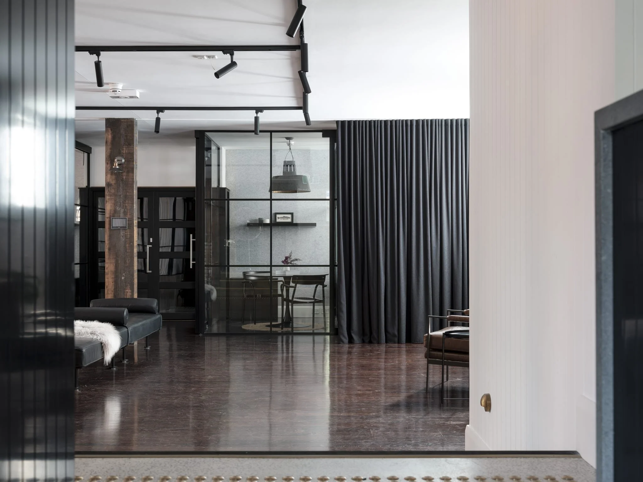 Modern interior space with dark wood flooring, black leather seating, black curtains, glass wall, and simple furniture.