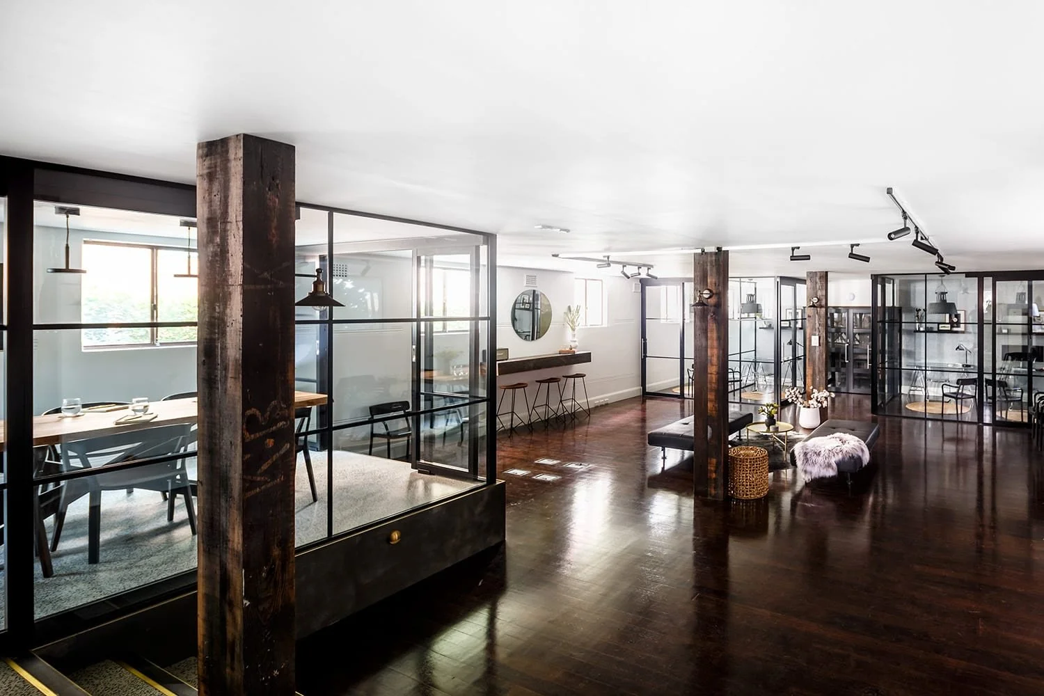 Modern open-plan office with glass-walled meeting rooms, wooden flooring, and minimalist decor including seating and tables.