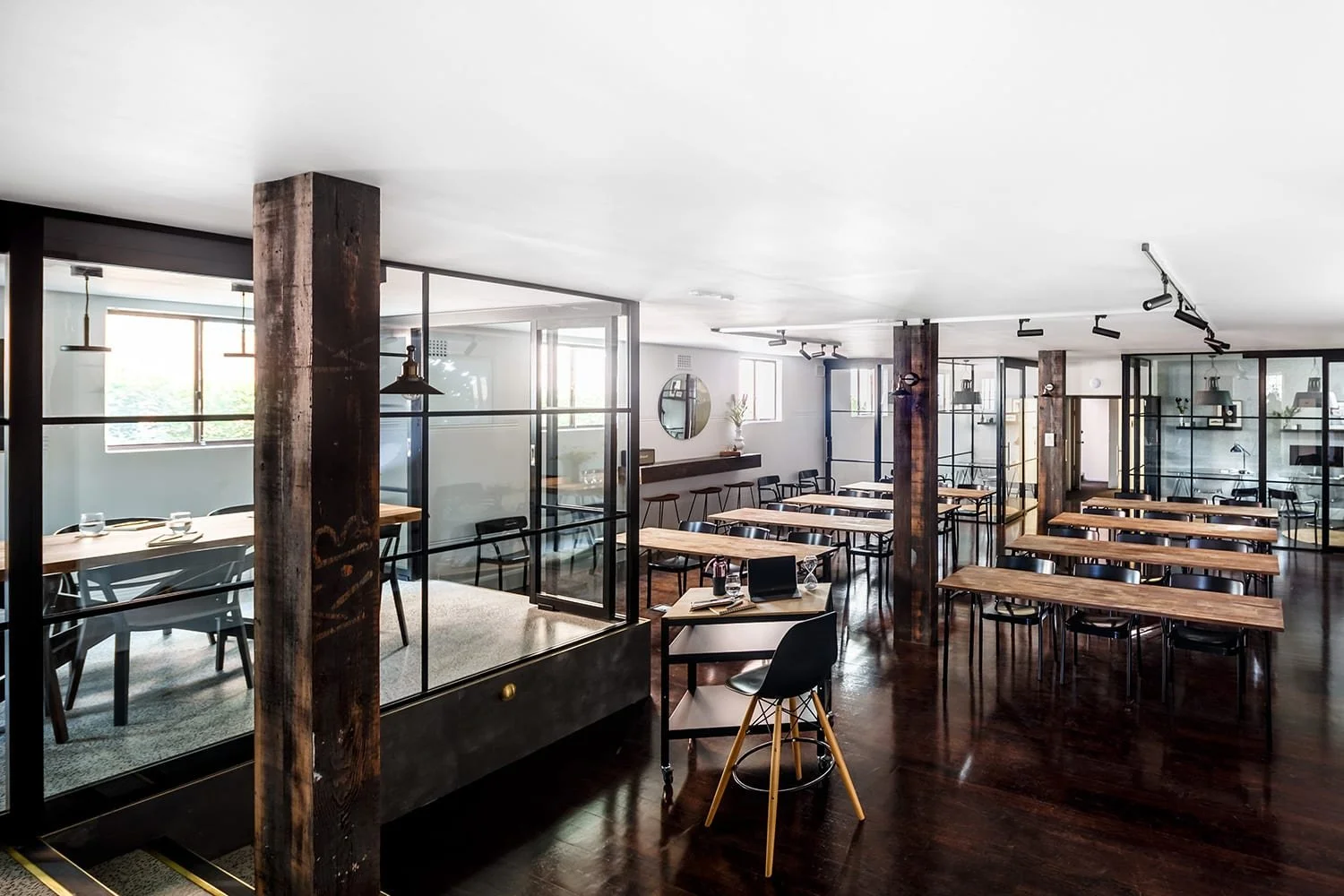 Modern restaurant interior with wooden tables, black chairs, glass partitions, and wooden support beams, illuminated by natural light from windows.