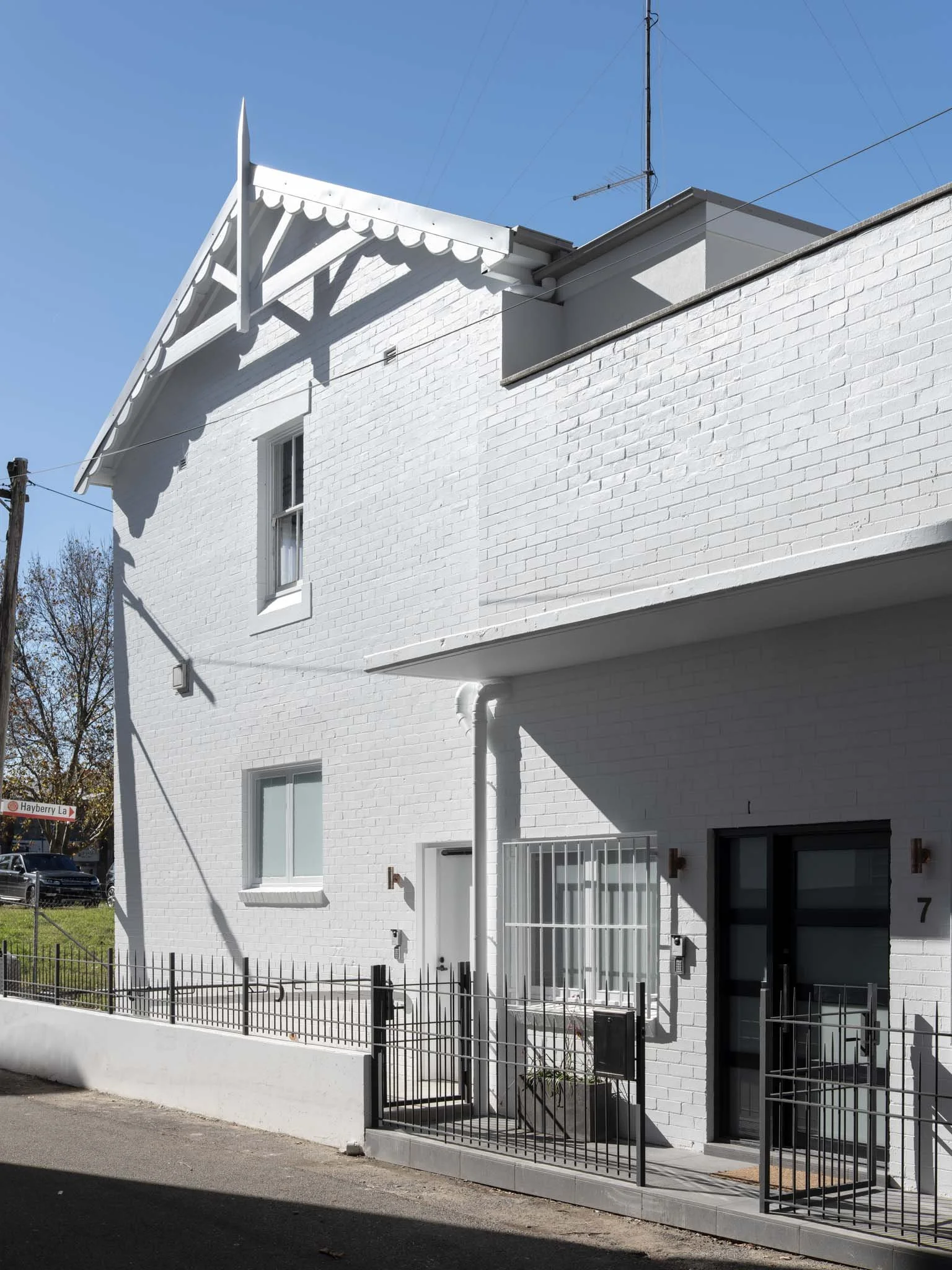 White brick residential building with black gate and small patio, multiple windows, and a decorative gable roof detail, under clear blue sky.