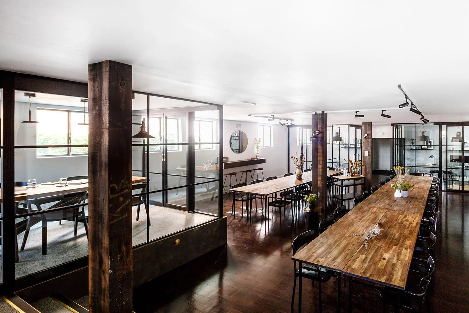 Modern dining area with large wooden tables, black chairs, and glass-enclosed sections, decorated with vases and plants.