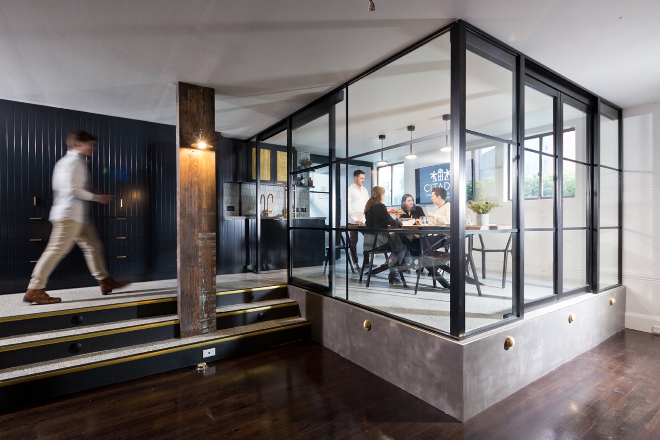 A modern office space with a glass meeting room and people working inside, seen from a hallway with stairs and a blurred person walking by.