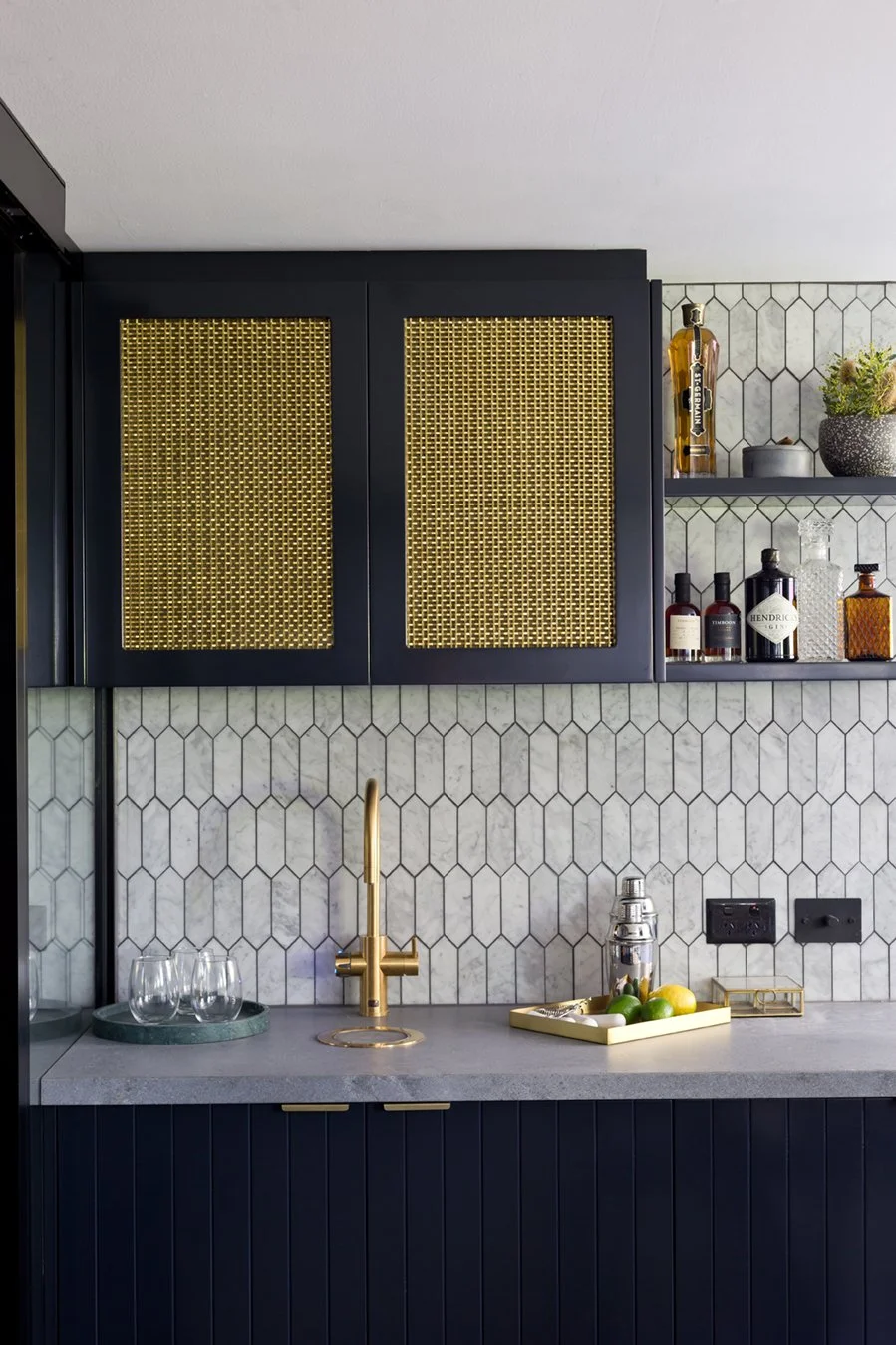 Modern kitchen with hexagonal tile backsplash, gold faucet, dark cabinetry with gold accents, open shelves with bottles and potted plants, and a small counter with glasses, fruit, and a citrus press.