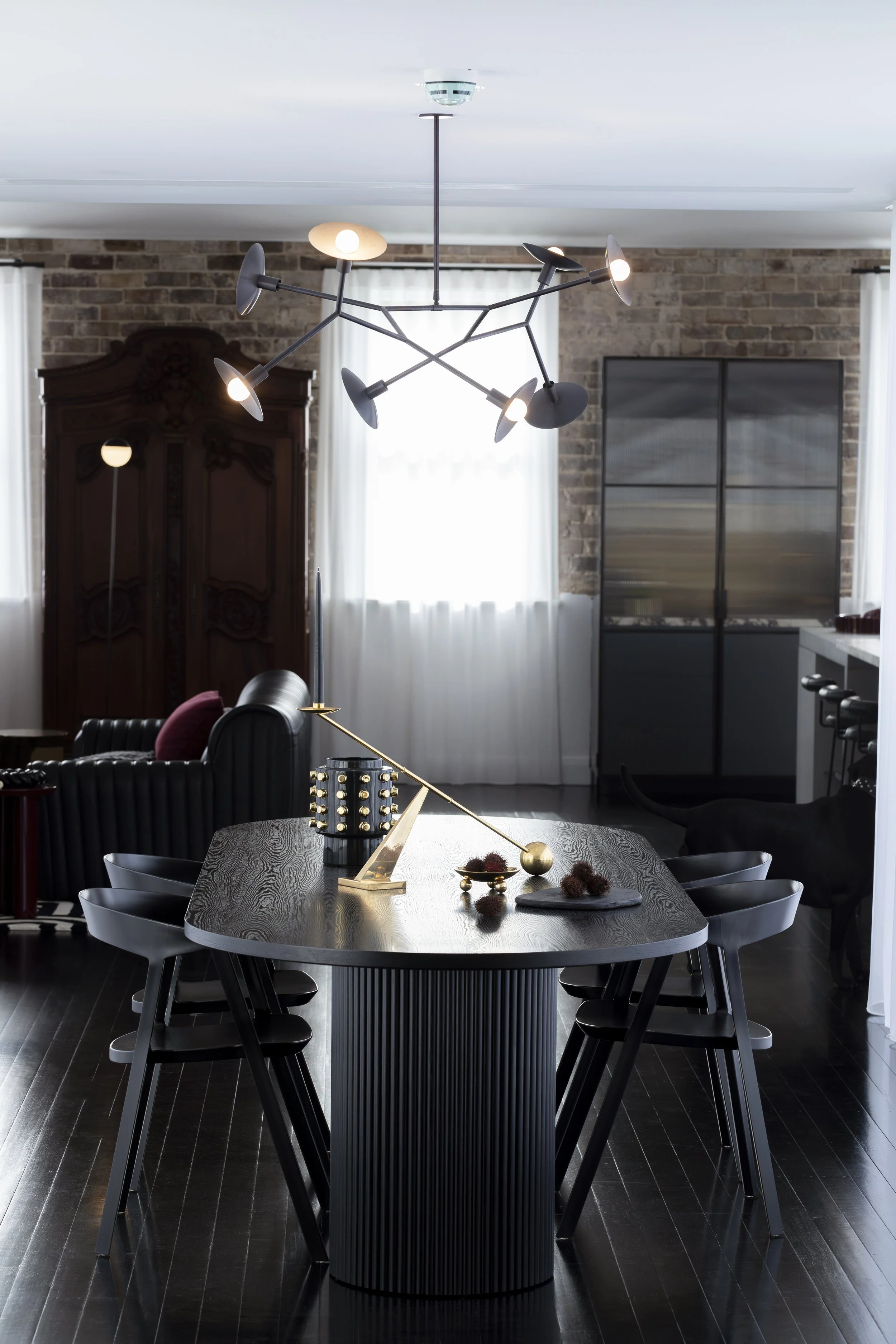 Modern dining room with black wood table, six black chairs, decorative gold and black desk lamp, black and gold decorative objects, abstract black chandelier, dark wooden floor, brick wall, white curtains, and a black dog near the kitchen area.