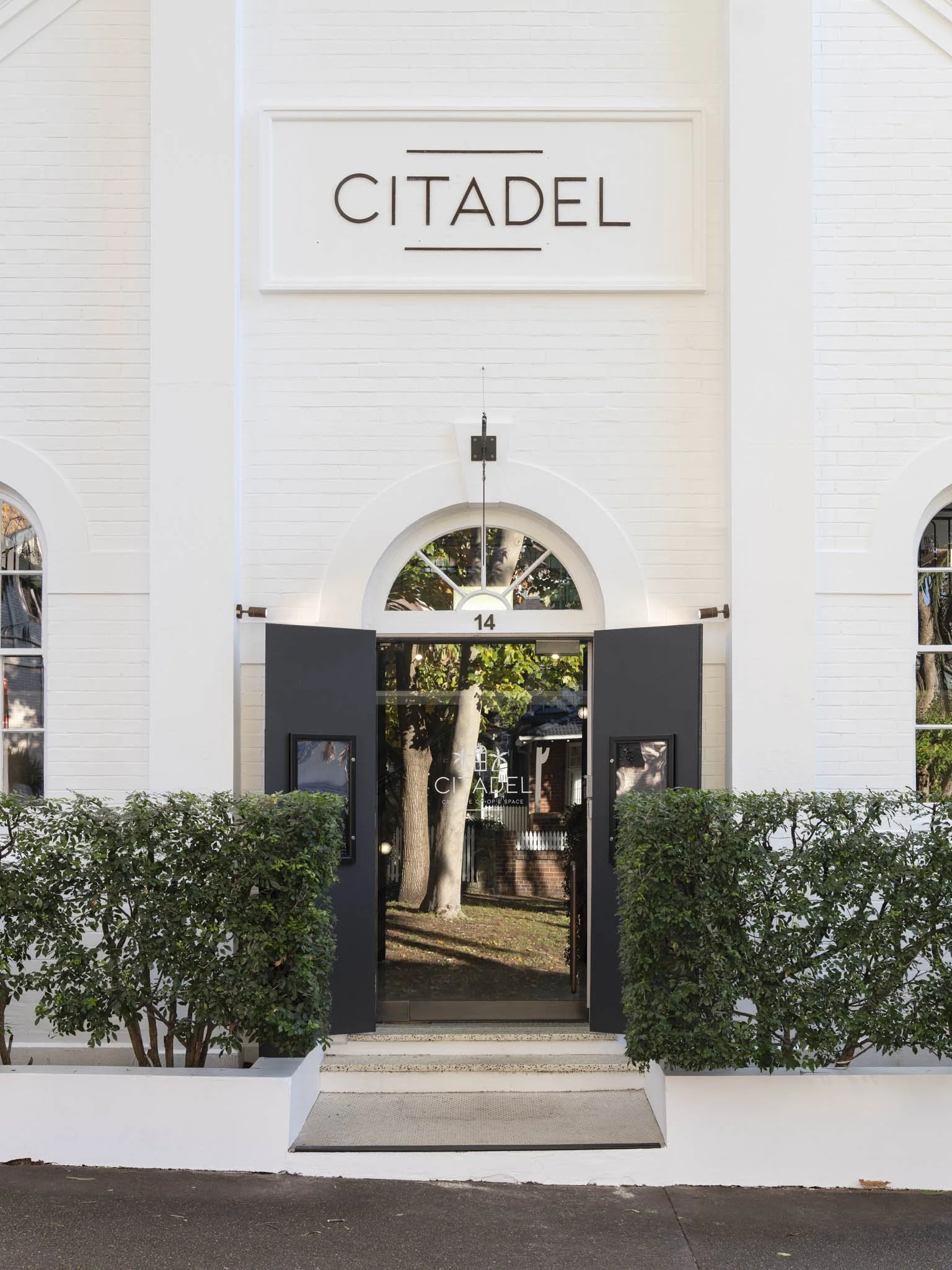 White building with the word CITADel on a sign above the entrance. The entrance has black doors that are open, revealing trees and greenery inside. There are bushes on either side of the entrance.