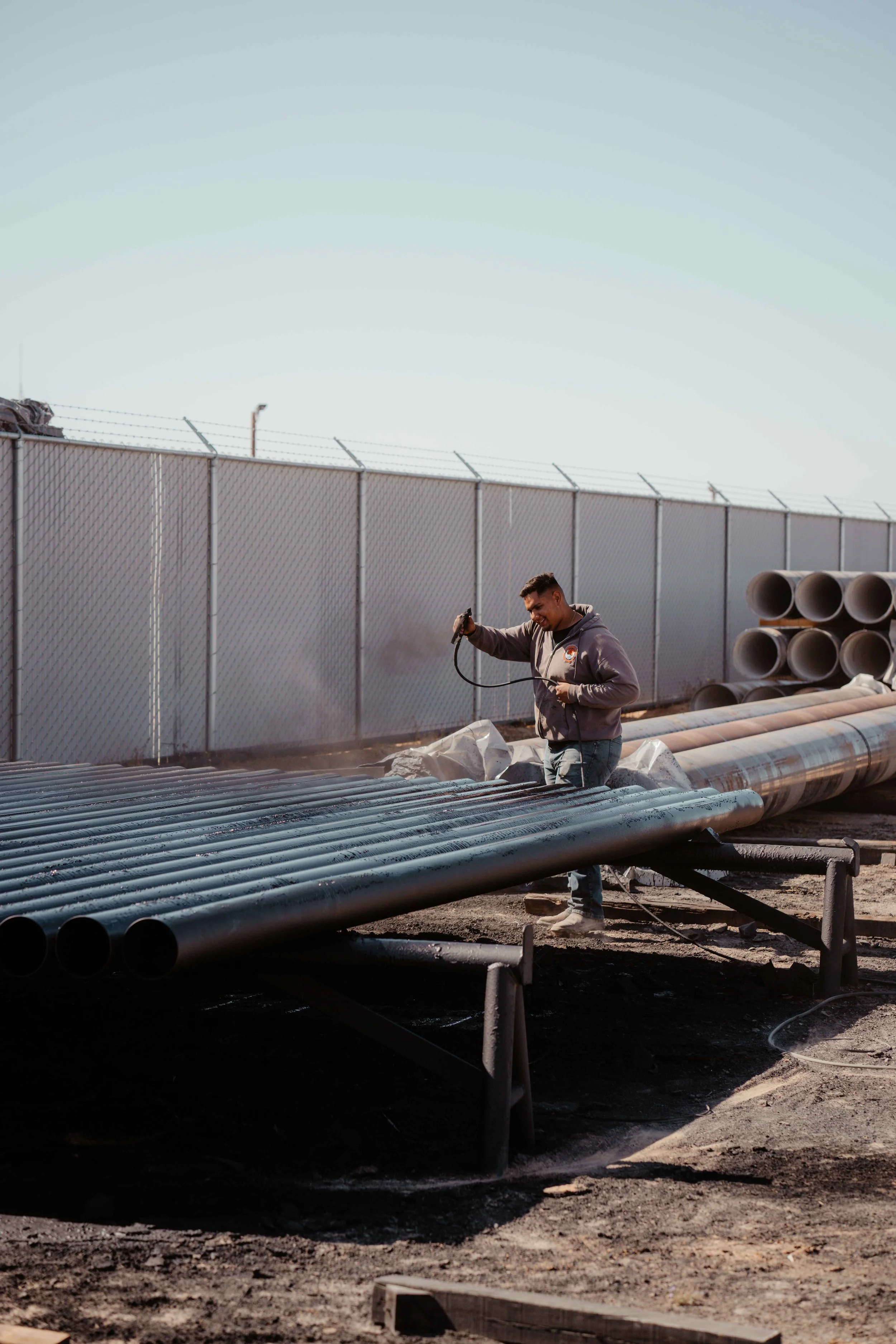 Man working outdoors with pipes, using spray equipment on a large pipeline near a fenced area.