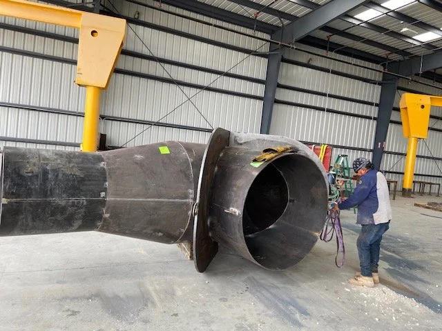 Person inspecting or working on a large industrial pipe or turbine inside a warehouse or factory.