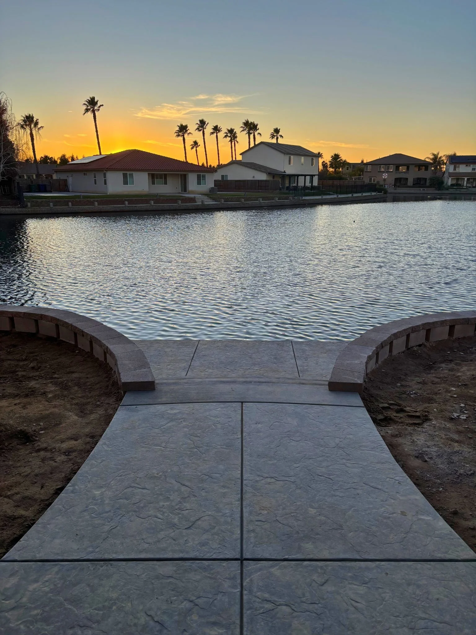 View of a canal or river at sunset, with houses and palm trees along the water and a paved walkway and curb in the foreground.