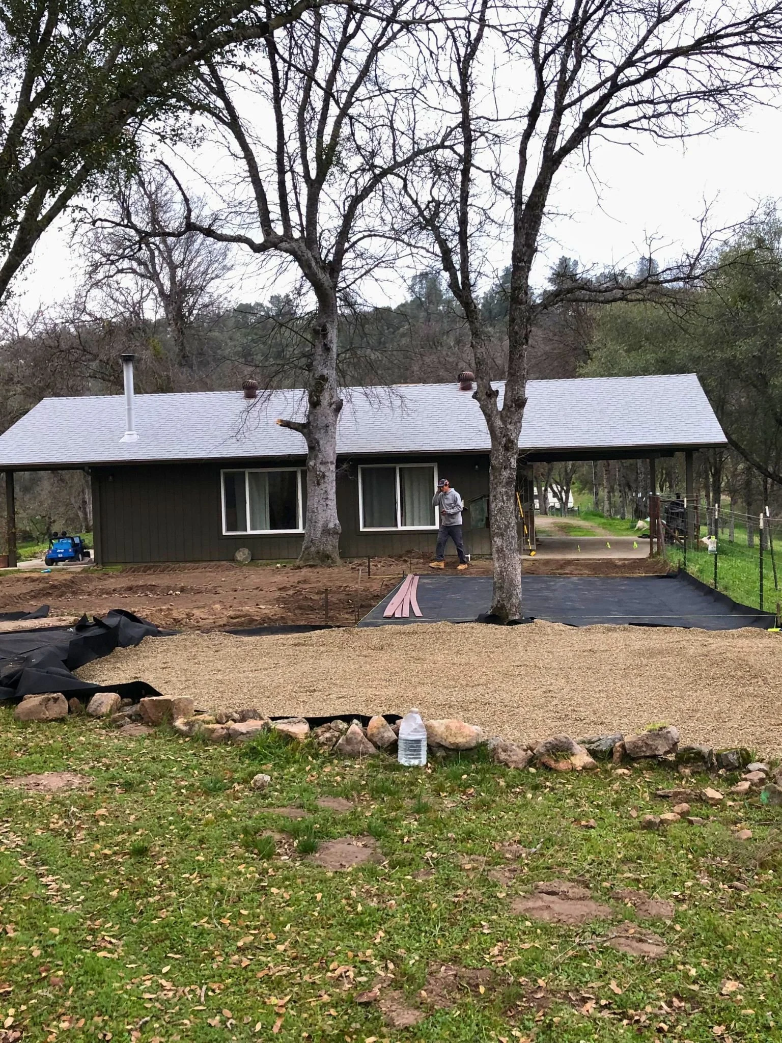 A backyard scene with a house, large leafless trees, a man standing and working, a large water bottle on the ground, and an area covered with gravel or mulch in the foreground.