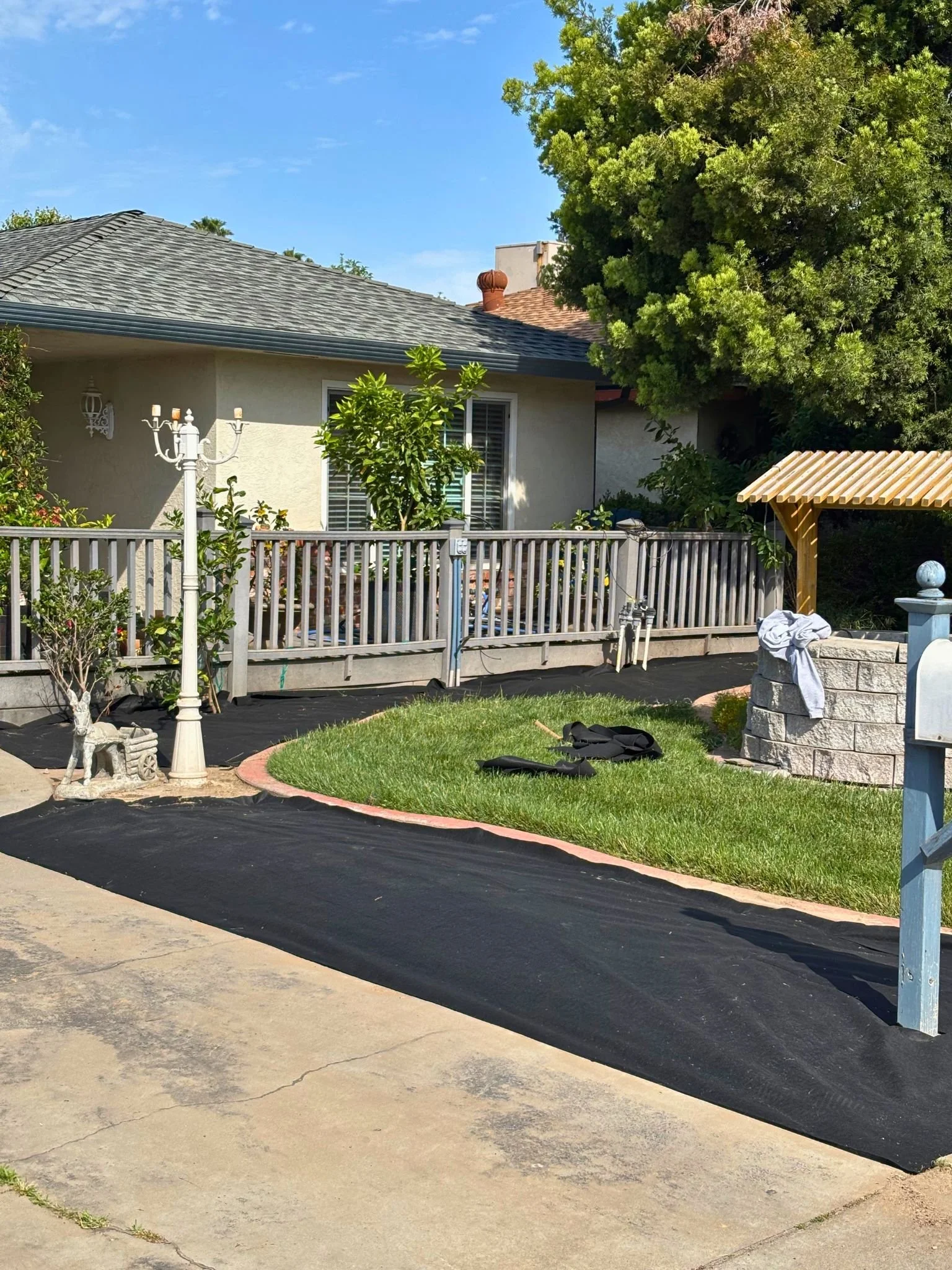 Backyard with a house, large tree, patio, and black landscaping fabric on the ground. There is a white fence, a lamp post, and some gardening materials and tools.