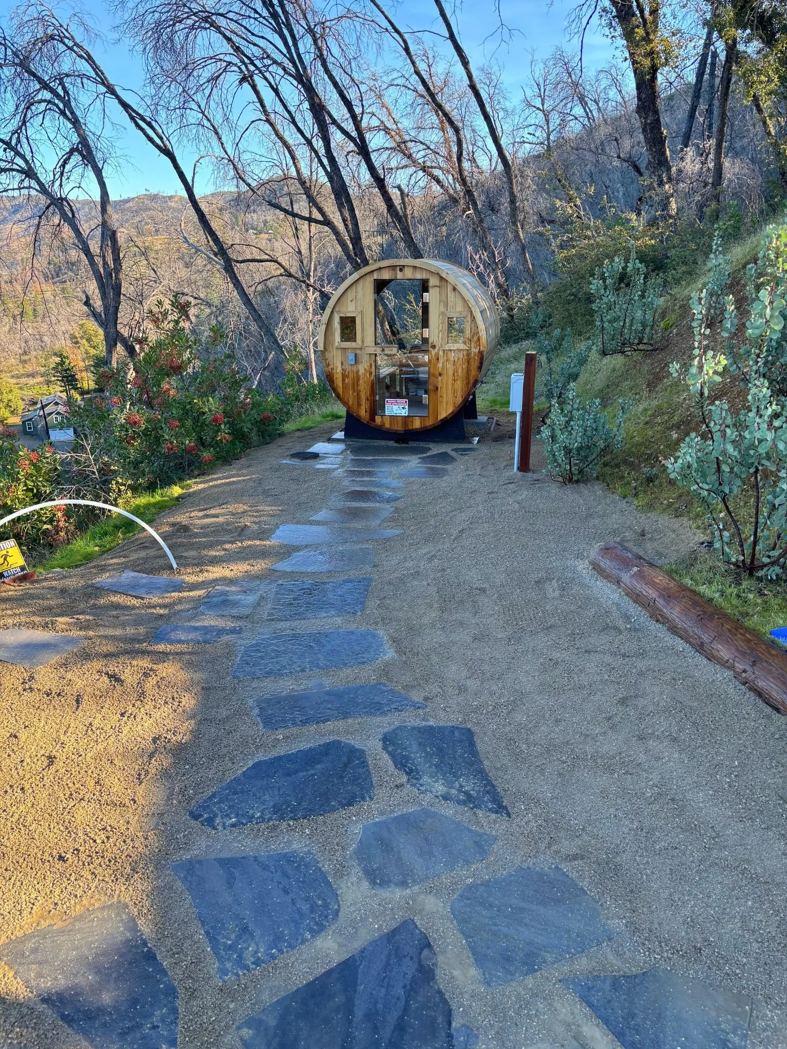 A wooden tiny house on wheels in a natural, mountainous setting with trees, bushes, and a gravel pathway.