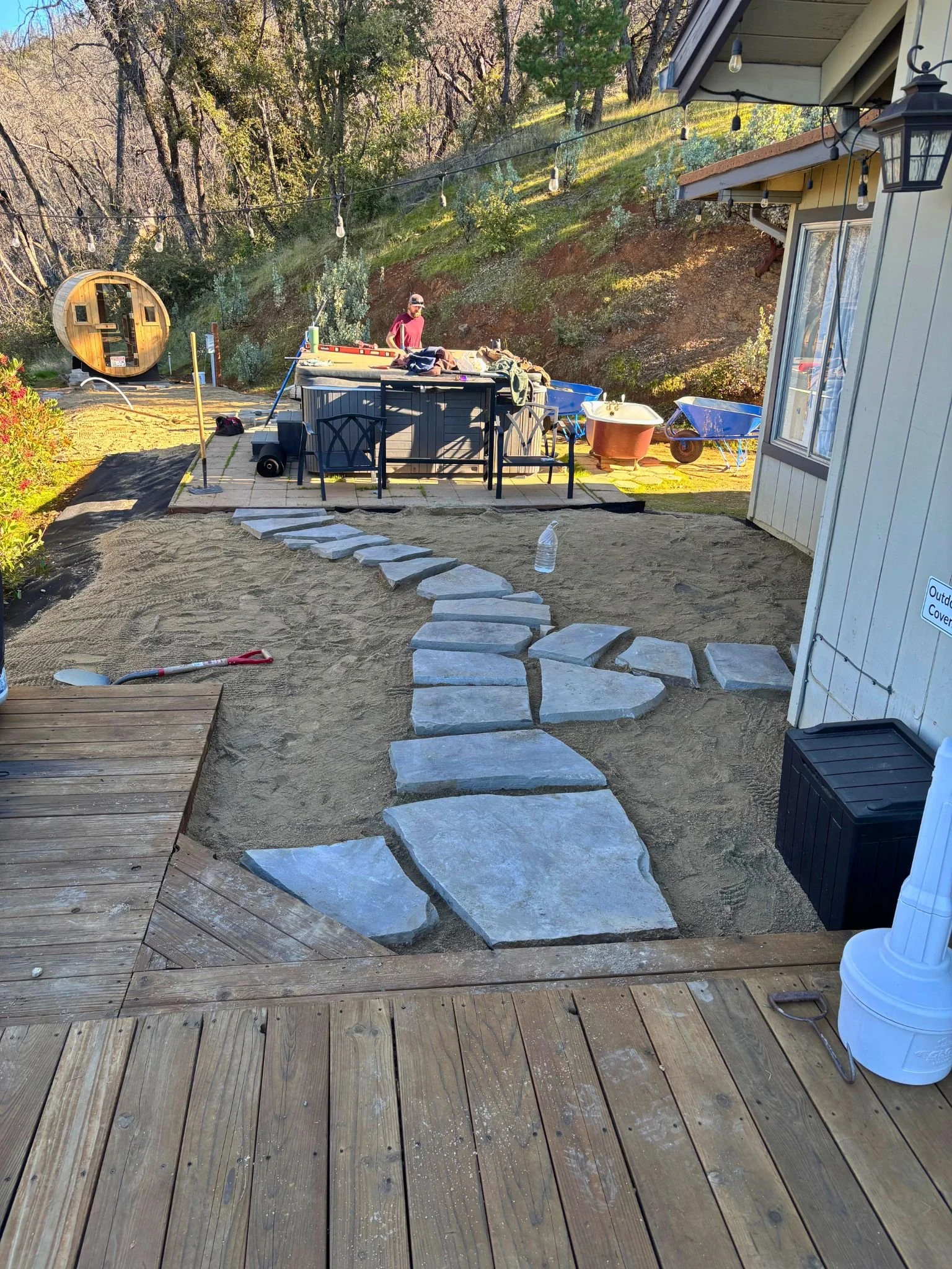 A backyard space with a wooden deck in the foreground, a stone pathway leading through the yard, a man working near a hot tub, a blue wheelbarrow, a bathtub, and a house with outdoor string lights. There are trees and a hillside in the background.