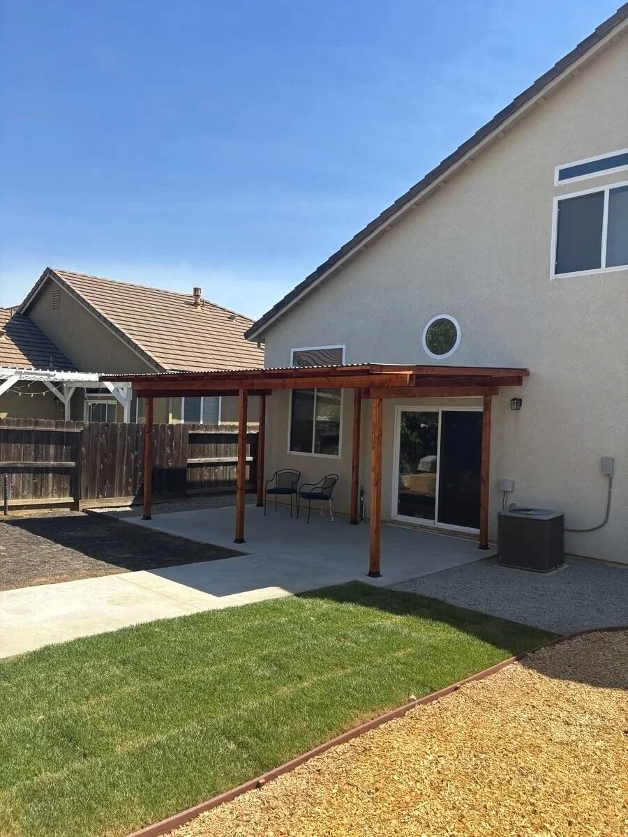 Backyard of a house with a wooden patio cover, chairs under it, and a sliding glass door. There is a grassy area, a concrete walkway, and a fenced yard with neighboring houses visible.