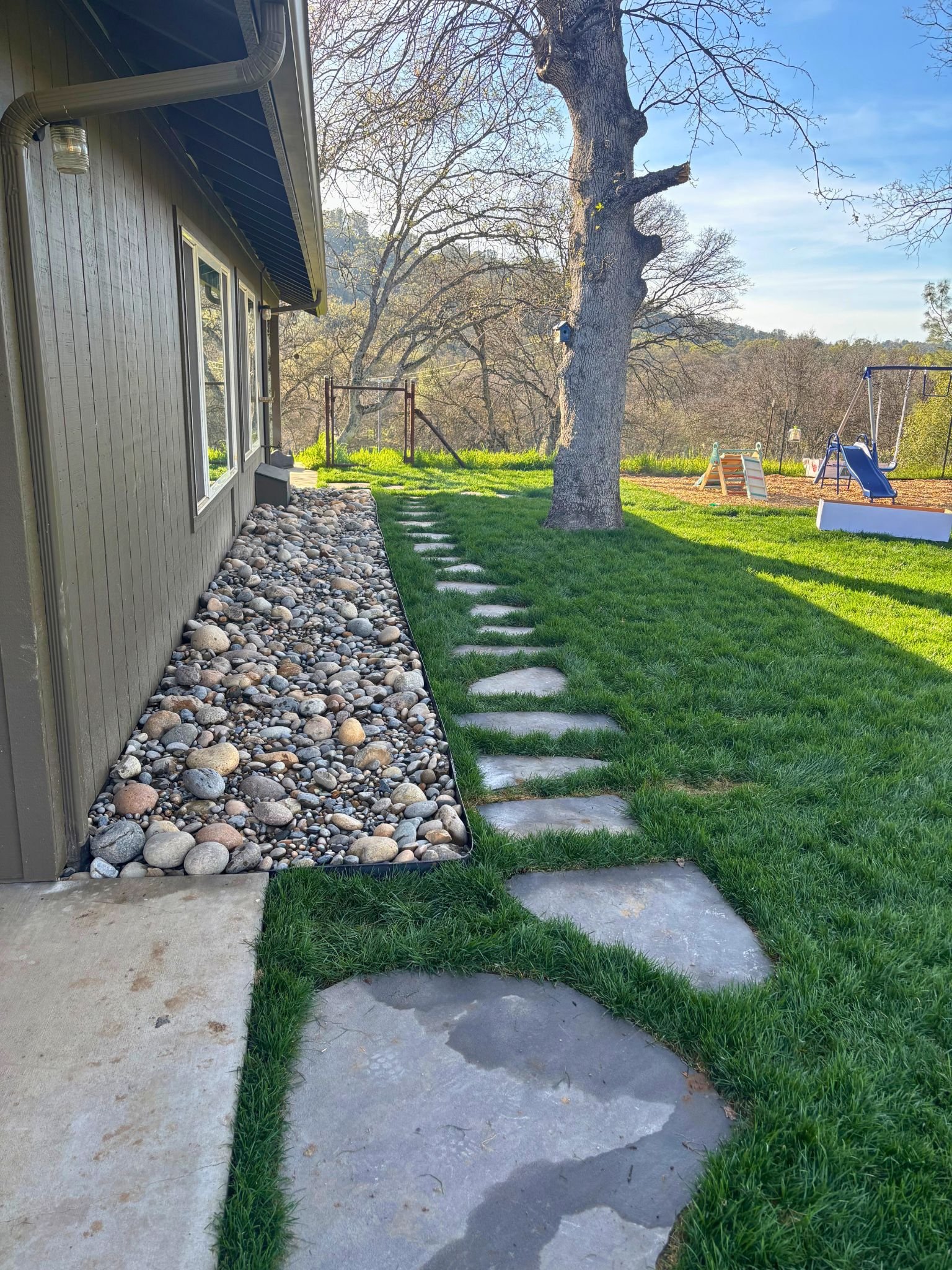 Side of house with grey siding, window, rock border, stepping stones, green grass, large tree, swingset, and a hill with trees in the background.
