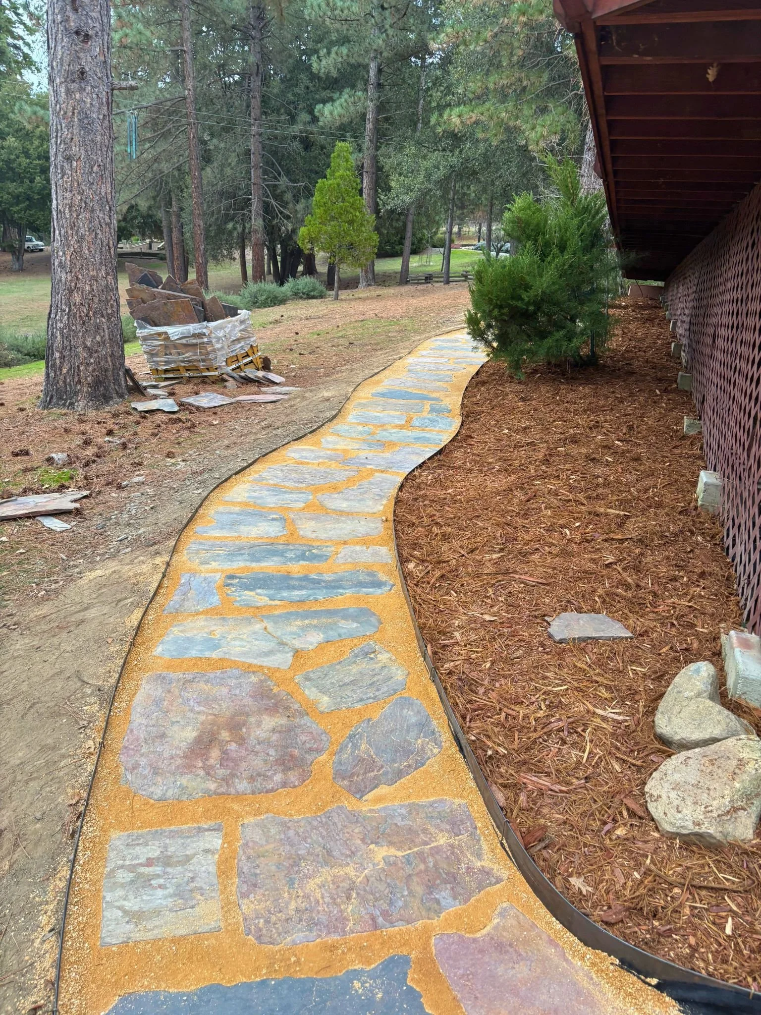 A winding stone path bordered by mulch and bushes in a wooded yard with trees, a house, and a stone wall in the background.