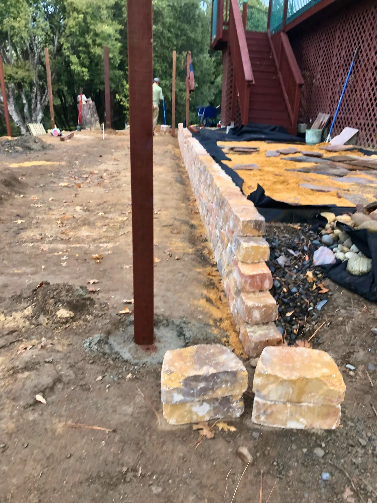 The image shows an outdoor construction site with a partially built stone wall, a black plastic sheet underneath, and a wooden staircase attached to a deck or house. There is a person working in the background, surrounded by trees and construction ma