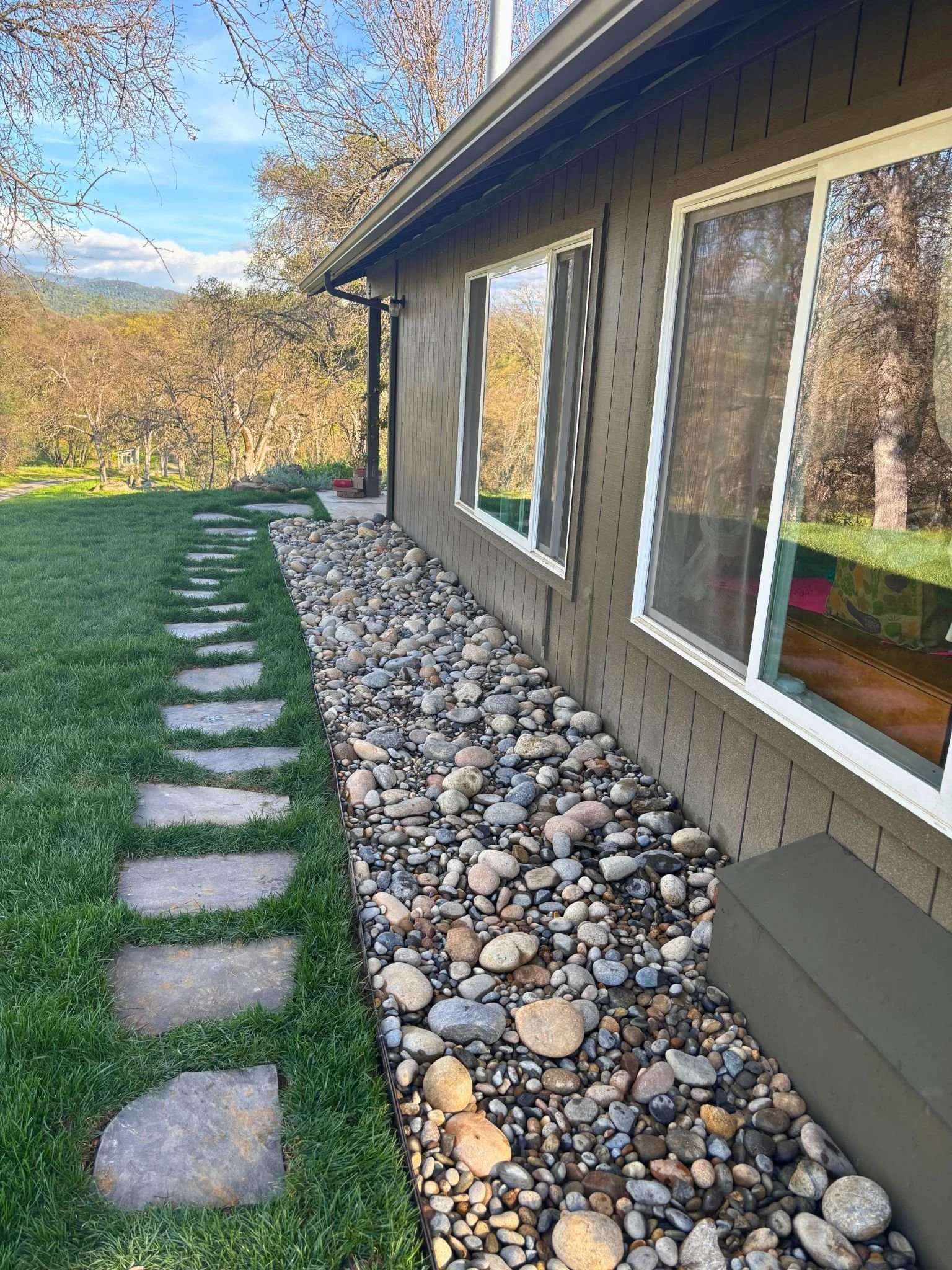 Side of a house with a gravel bed, stone pathway, and large windows, overlooking a grassy yard with trees and hills in the background.