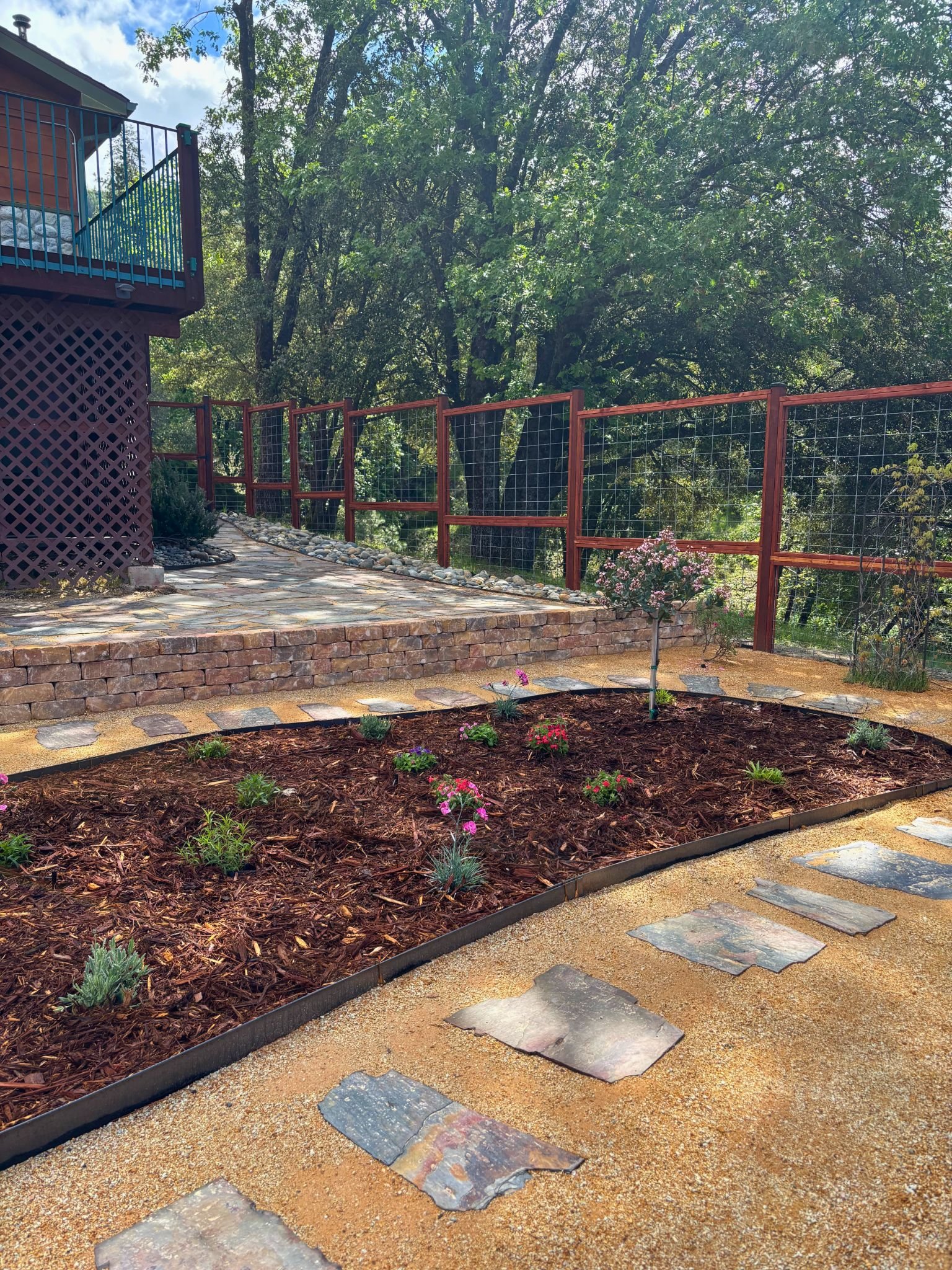A backyard garden with landscaped flower beds, a stone pathway, a wooden deck with railing, trees in the background, and a fence.