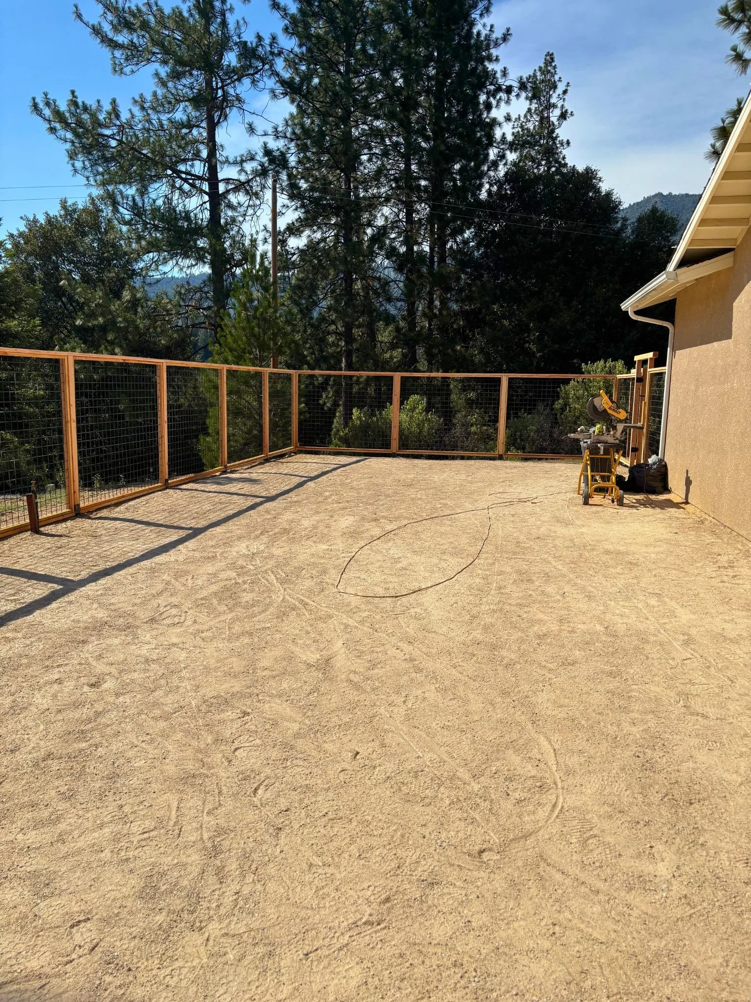 An outdoor patio area with a sandy or dirt ground, wooden fence, and tools leaning against the house wall, surrounded by tall pine trees and a mountainous landscape in the background.