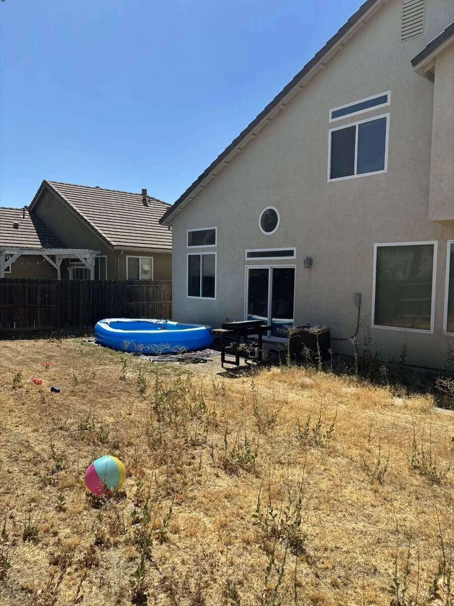 Backyard with a blue inflatable pool, a small black table, and a pool skimmer. The yard has dry grass and weeds, with a colorful beach ball in the foreground. The house is beige with white window trims, and a wooden fence surrounds the yard. Other houses are visible in the background under a clear blue sky.