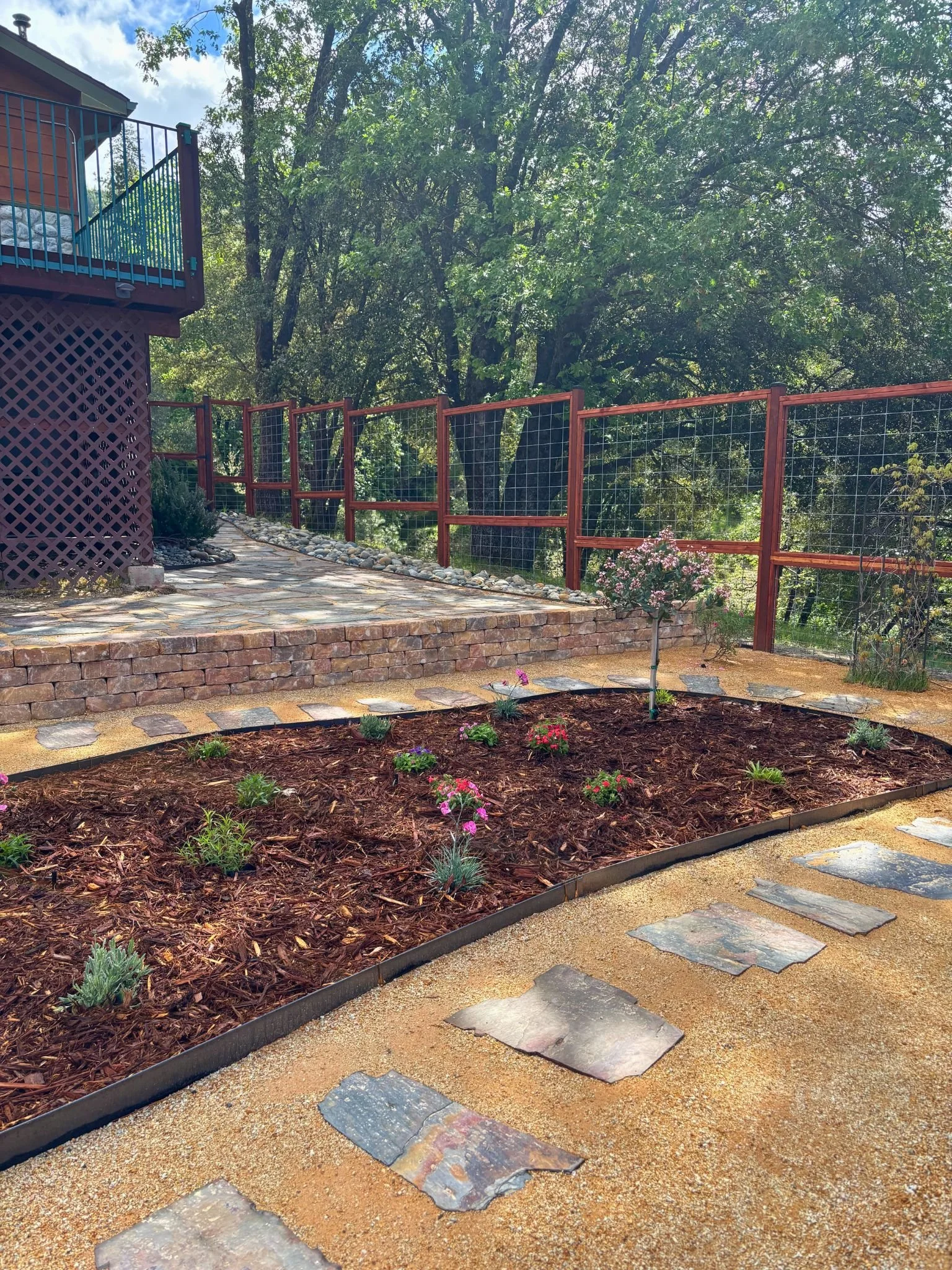 A backyard garden with a brick and stone pathway, small flowering plants, a raised flower bed with mulch, a red metal fence, and a tall tree with green leaves surrounded by other trees and greenery.
