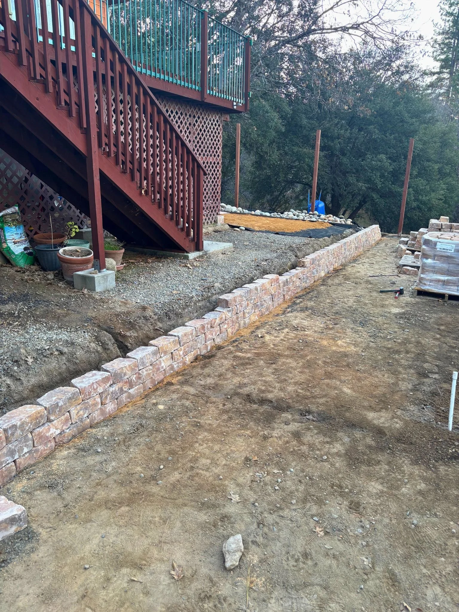 A backyard outdoor area under construction with a partially built brick border, a staircase leading to a deck, and construction supplies. The ground is dirt with rocks scattered around.