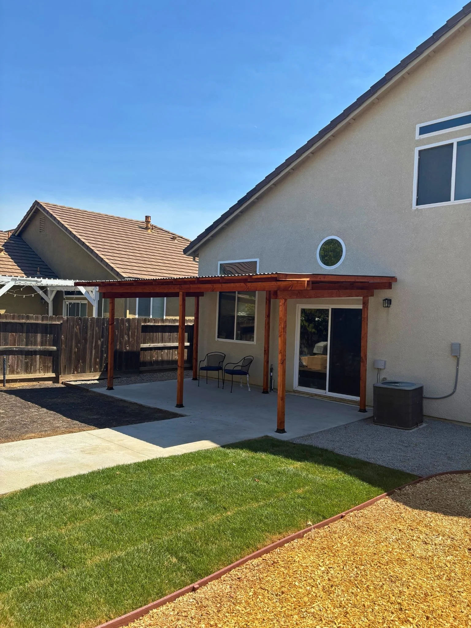 Backyard patio area with wooden pergola, chairs, and a sliding glass door on a beige house. Green lawn and decorative gravel border in the yard, with neighboring house roofs visible under a clear blue sky.