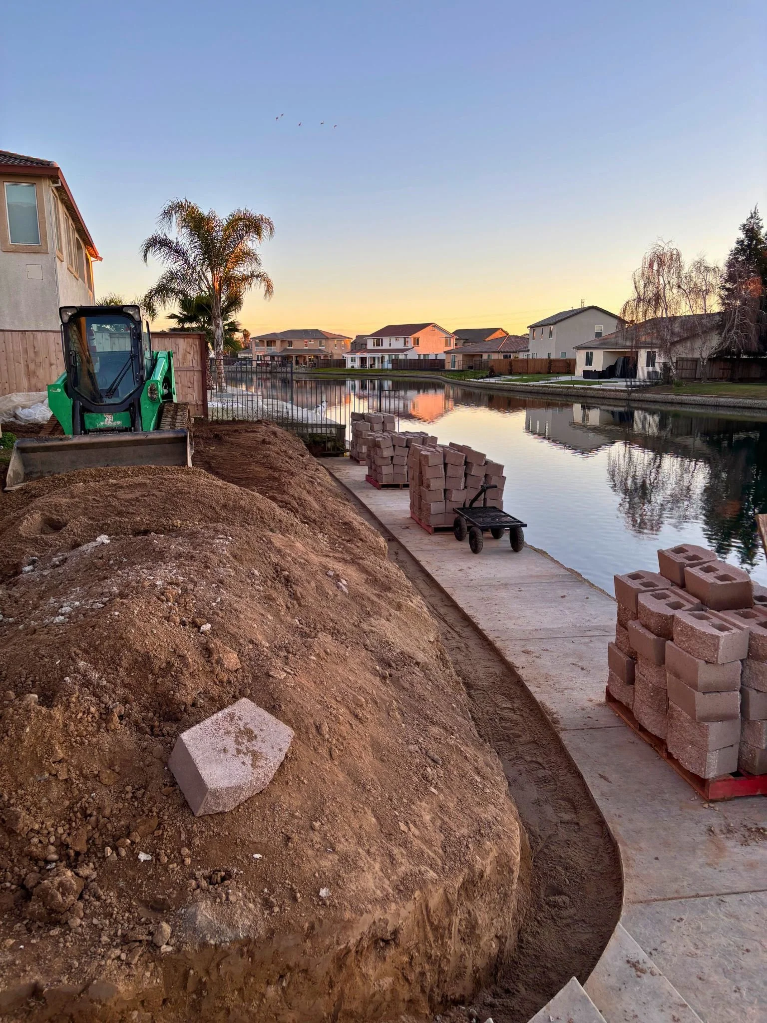 Construction site near a waterway with a small green excavator, piles of bricks, and a pathway under construction in a suburban neighborhood at sunset.