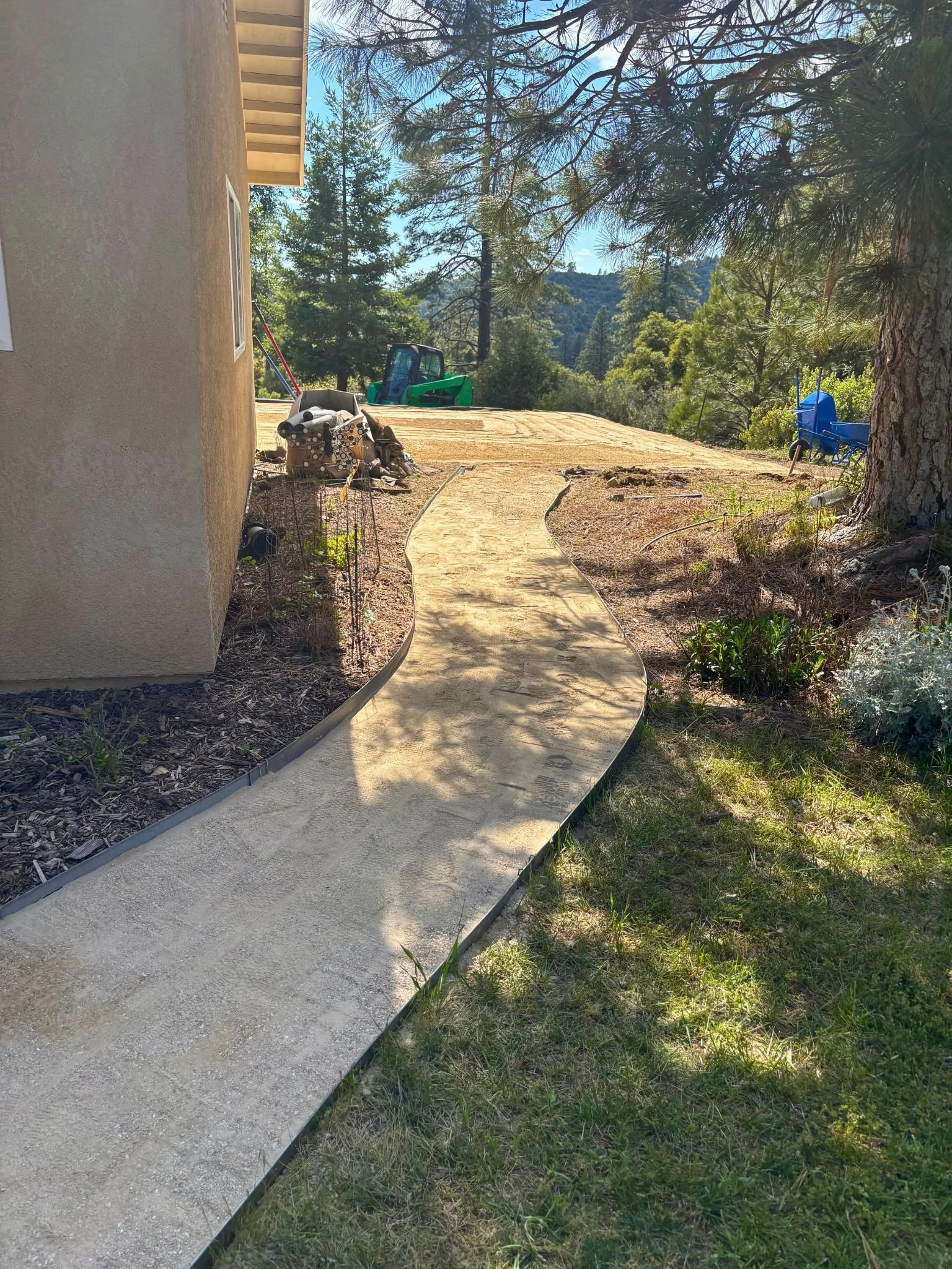 A newly laid concrete walkway curves around the side of a house, supported by a metal frame. The house wall on the left side has a small window and beige stucco paint. In the background, construction materials and equipment, including a small green t