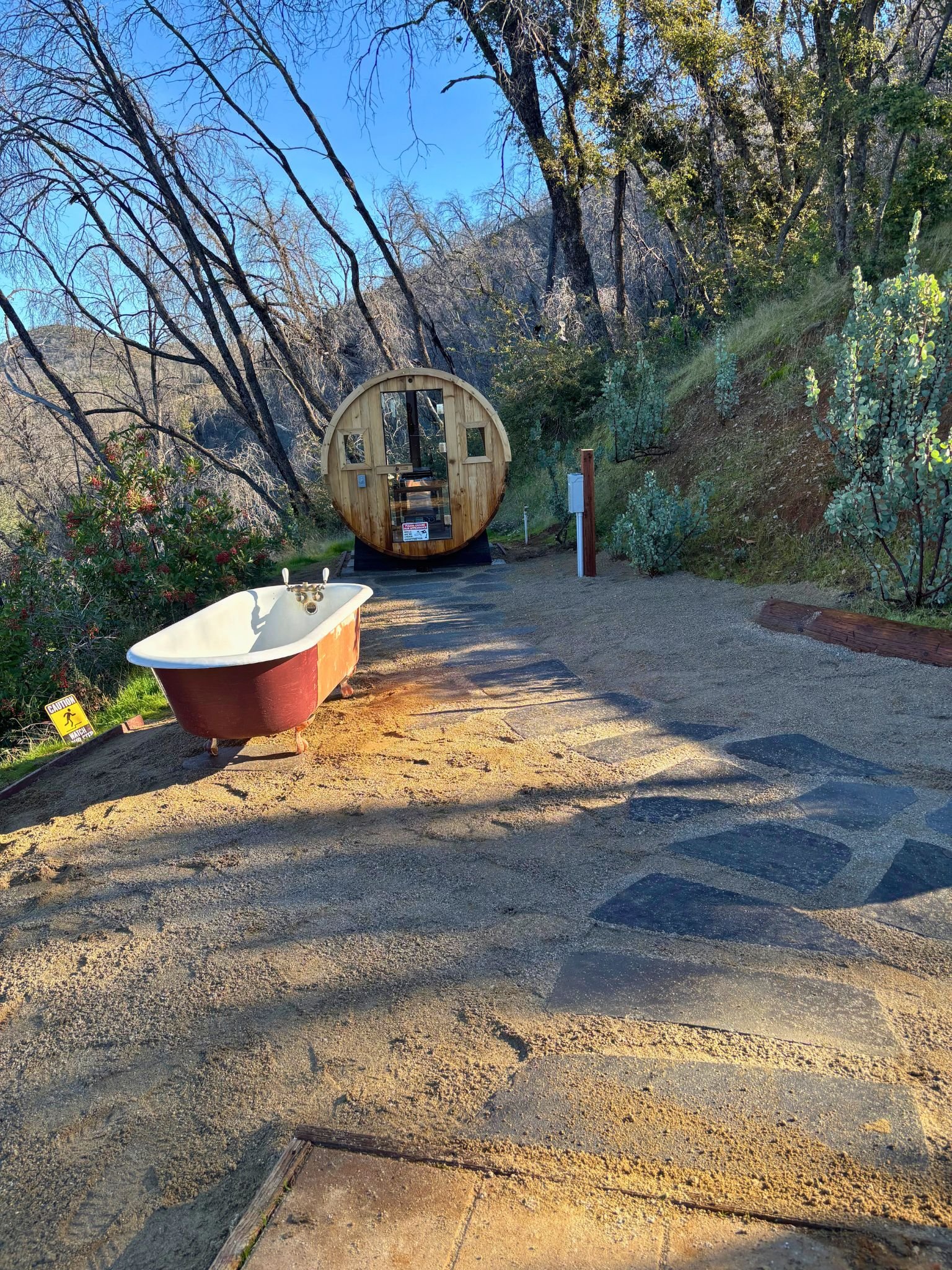 Outdoor scene showing a dirt trail with a vintage bathtub, a small cabin, and leafless trees against a clear blue sky.