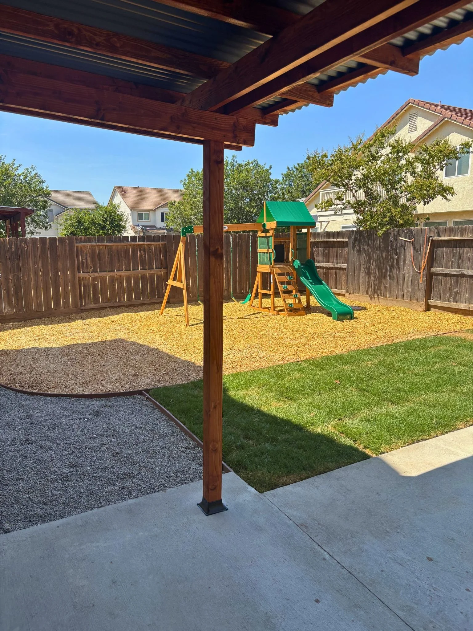 Backyard with a wooden fence, a play structure with a slide and swings, a grass lawn, and neighboring houses with trees in the background.