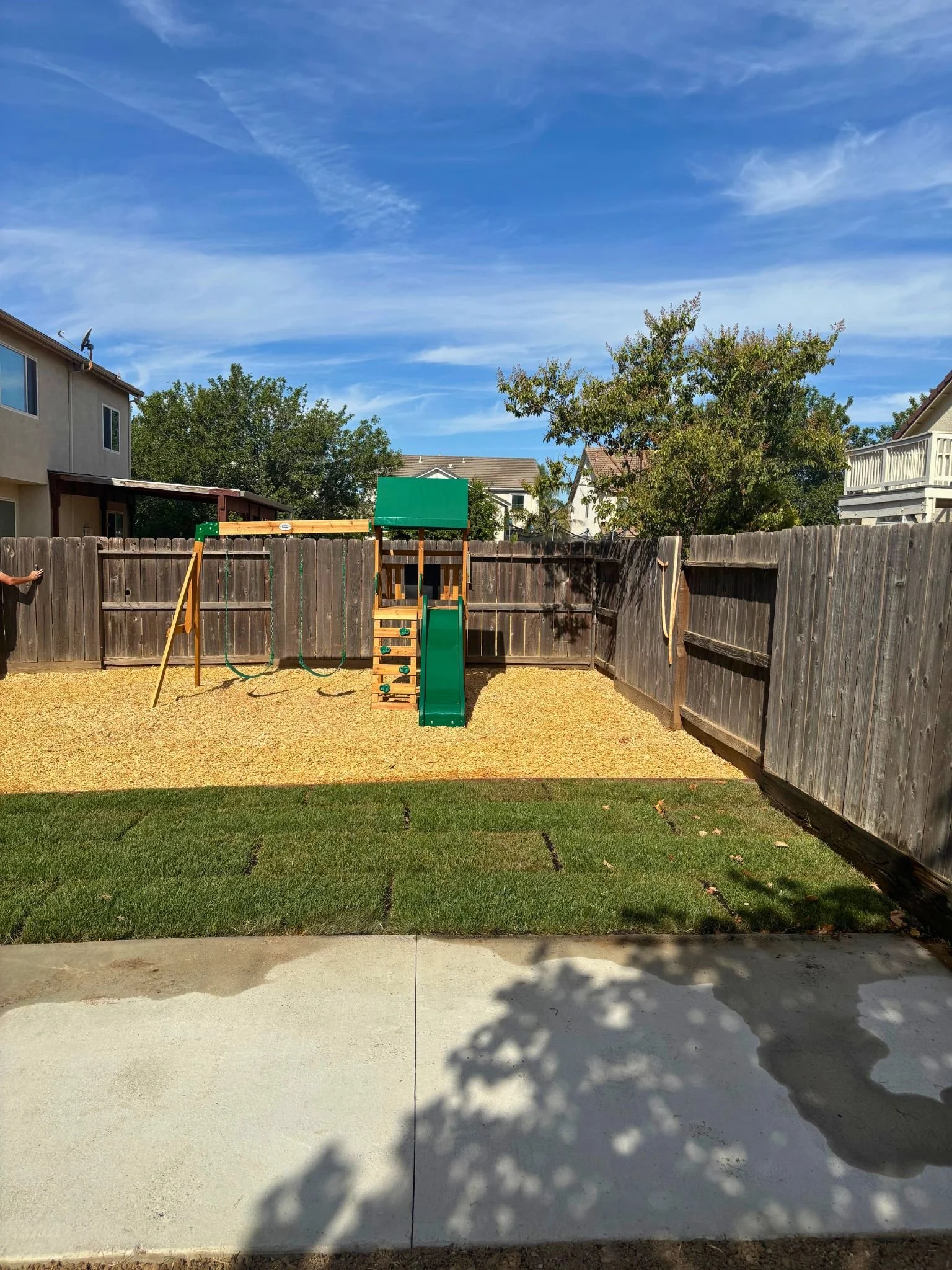 A backyard with a wooden fence, a small green slide, and a swing set, with a tree and neighboring houses under a blue sky with wispy clouds.