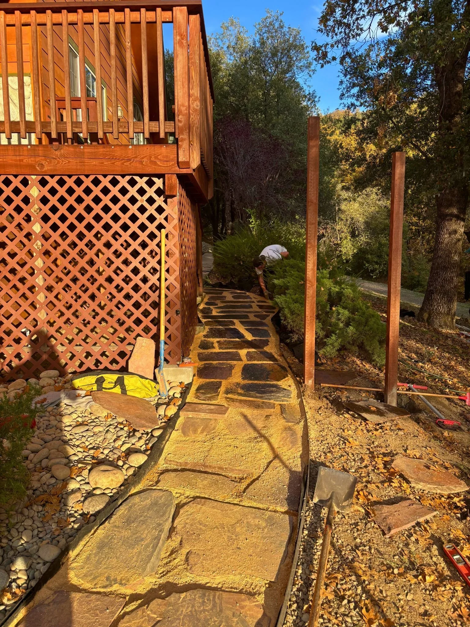 A person working on a landscaping project outside a wooden house, installing a stone pathway bordered by rocks and sand amid trees and a clear sky.