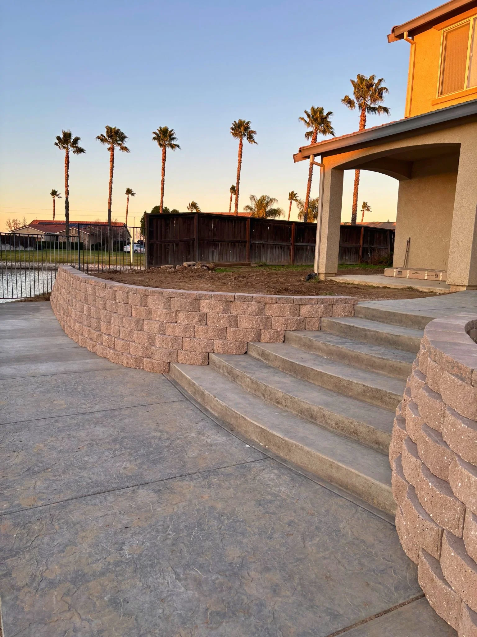 Back patio area of a house with steps leading down to a concrete surface, a curved brick retaining wall, and a partially finished backyard with a dirt area, surrounding wooden fence, and tall palm trees at sunset.