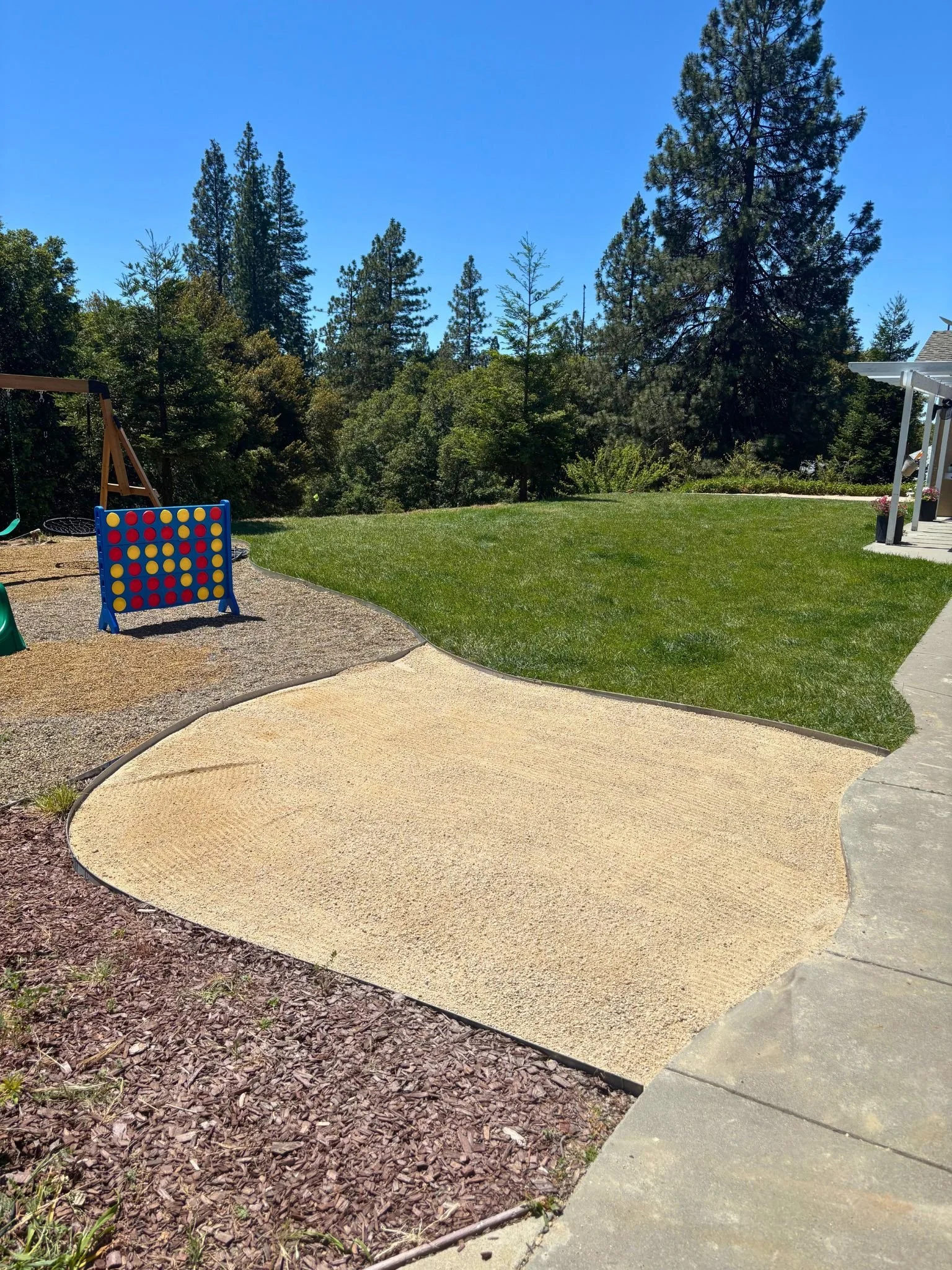 A backyard with a swing set on the left, a gravel area with a large connect four game, lush green grass, tall trees in the background, and a concrete path on the right.
