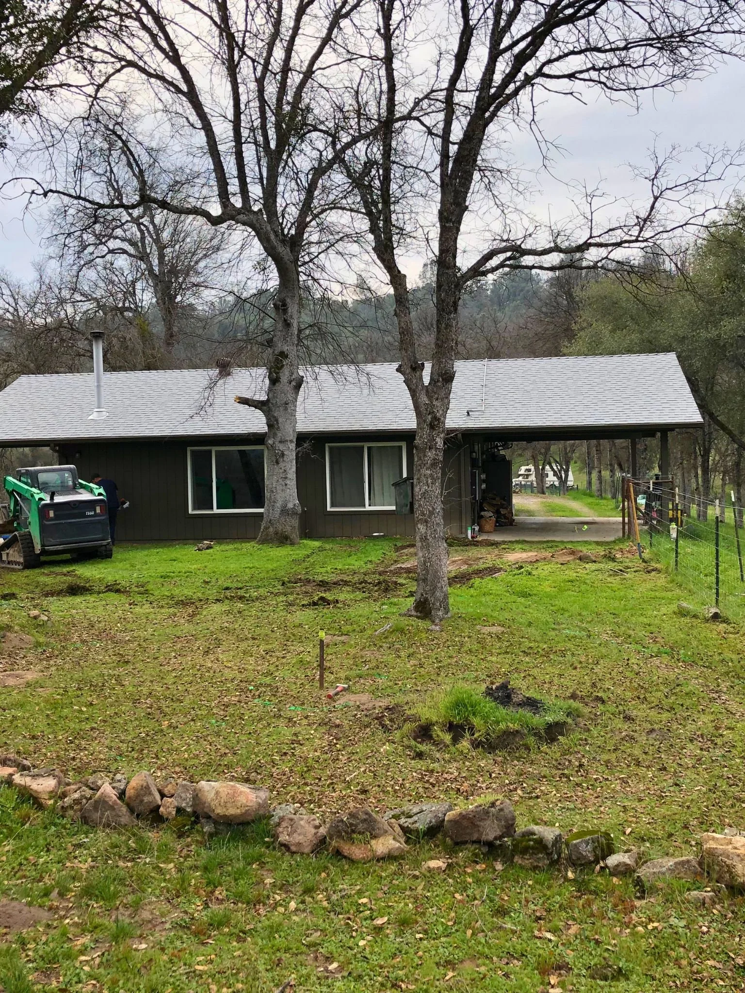 Backyard scene with two leafless trees, a house with a gray roof, and a green lawn with scattered rocks and some construction tools or equipment.