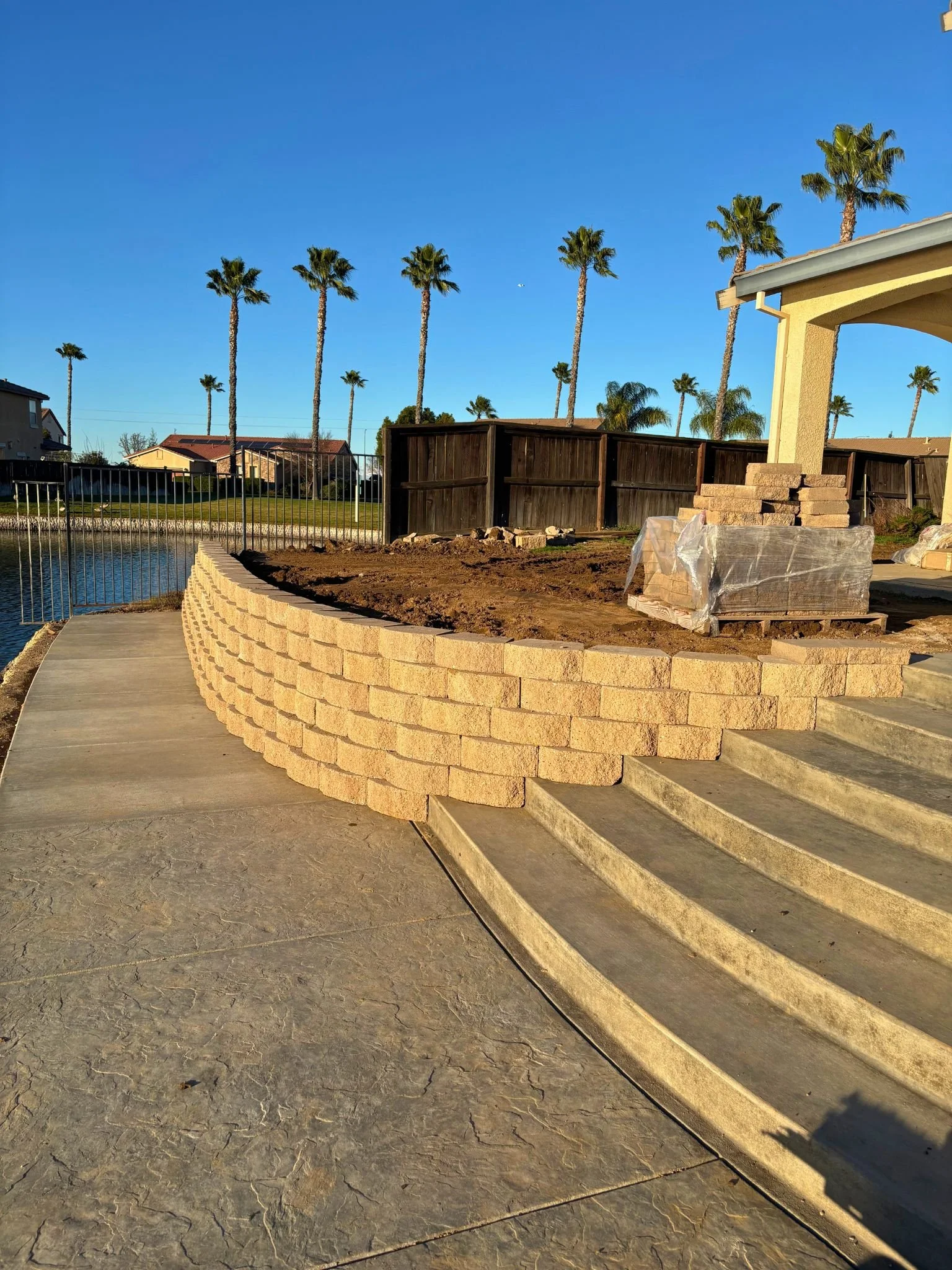 Backyard with tall palm trees, a wooden fence, a small pond, and a partially constructed brick wall on a sunny day.